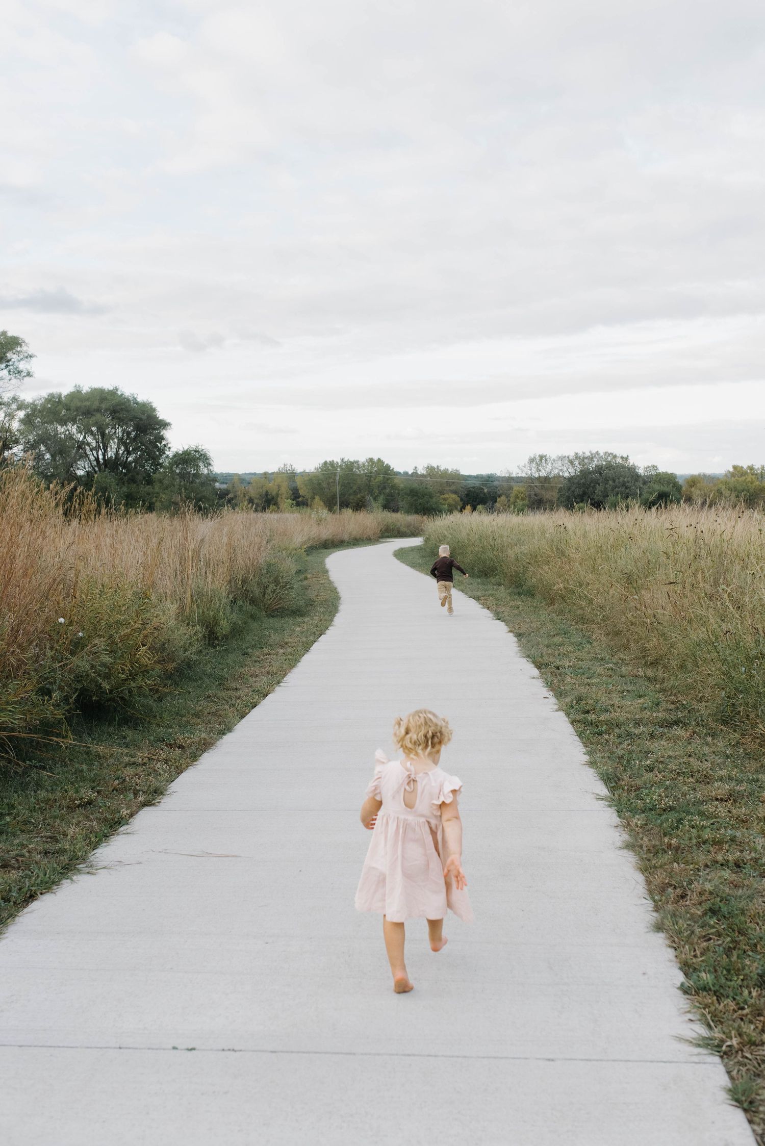Two young children walking down a long winding path flanked by tall golden grass under cloudy skies.
