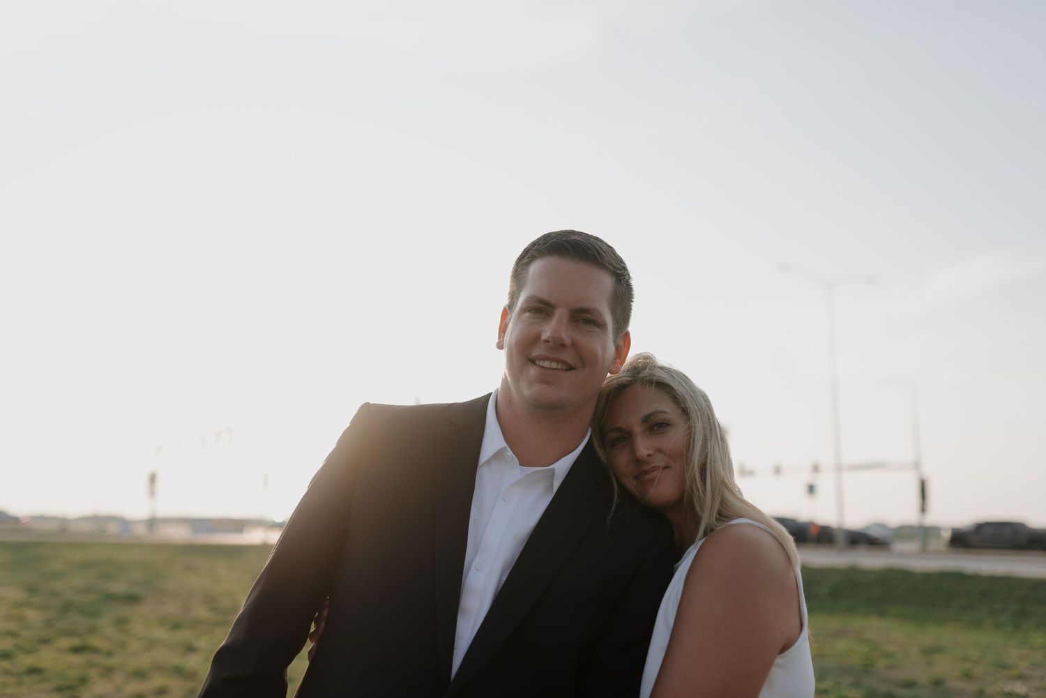 Engaged couple smiling together outdoors with soft backlit sunset glow behind them.