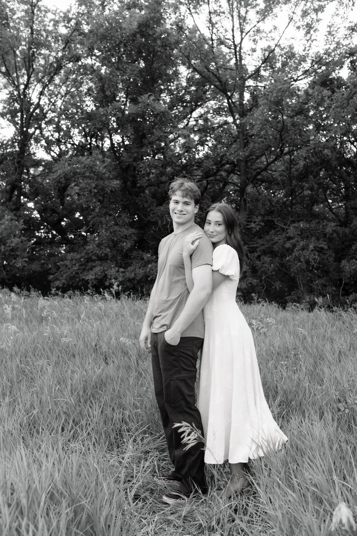 Black and white photo of a young couple posing in a grassy field with trees in the background.