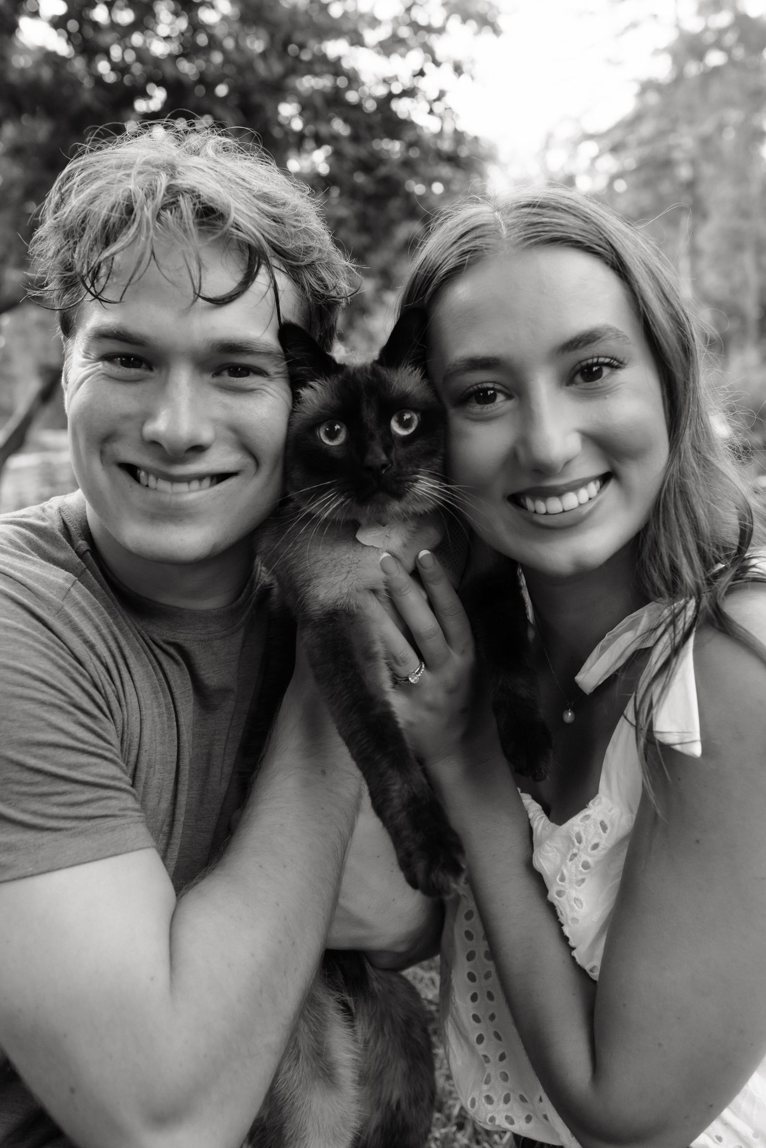 Black and white photo of a smiling young couple holding a dark Siamese cat outdoors.