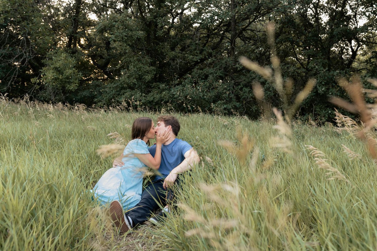 Couple kissing romantically in a lush green meadow surrounded by tall grass and trees at dusk.