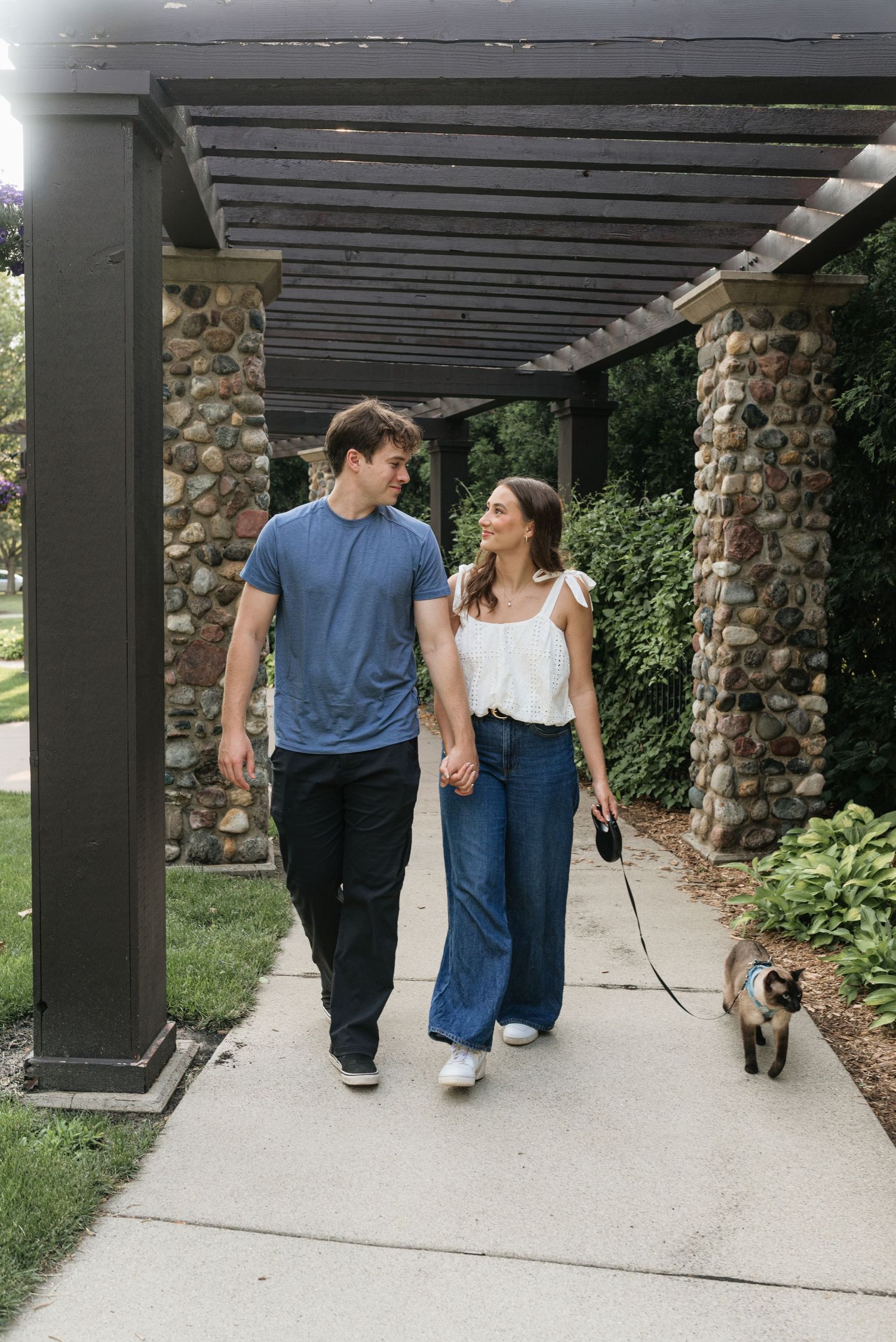 Smiling couple holding hands strolling under stone pergola while walking their dog on a leash.