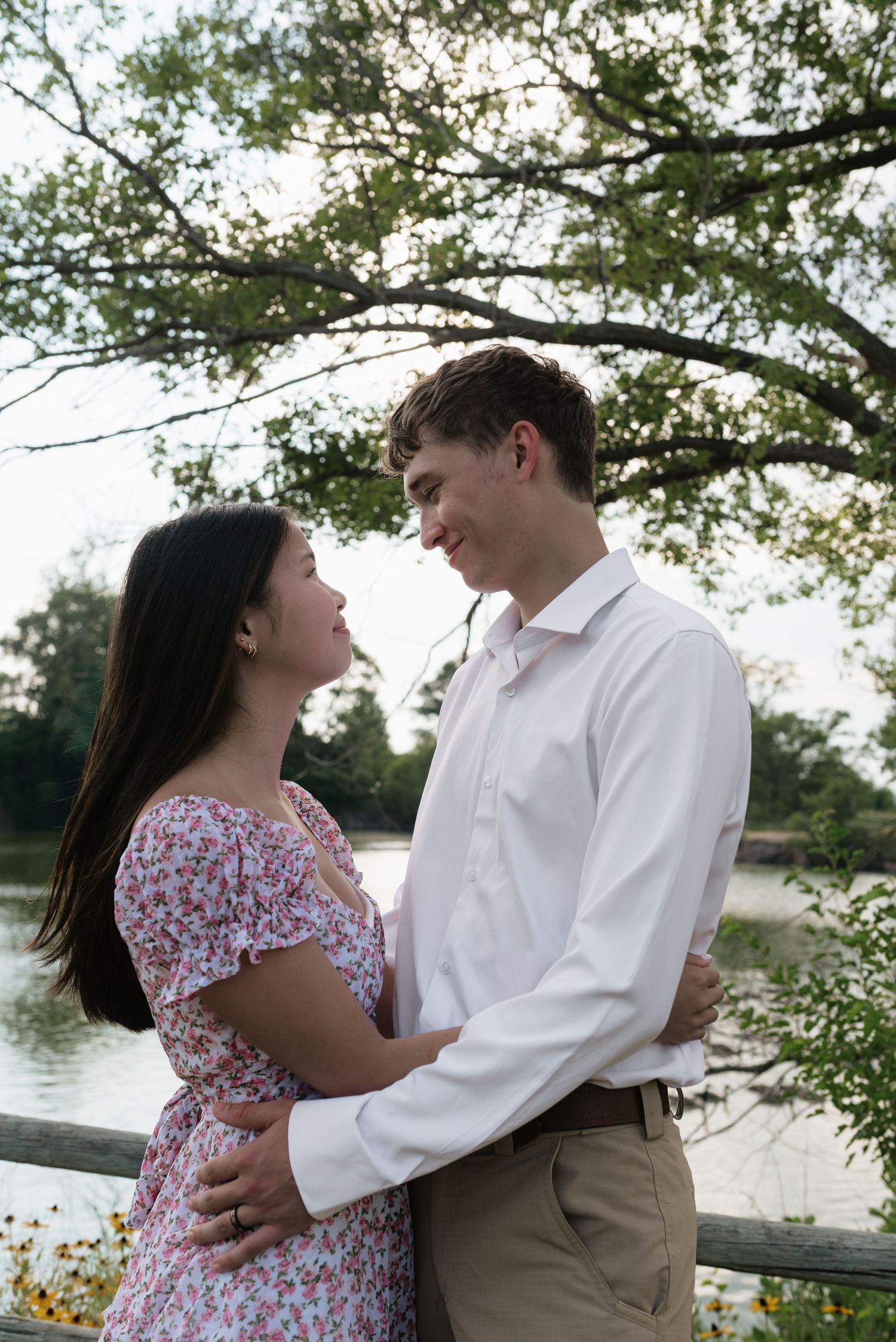 Couple embracing and smiling at each other near a lake, woman in floral dress and man in white shirt.