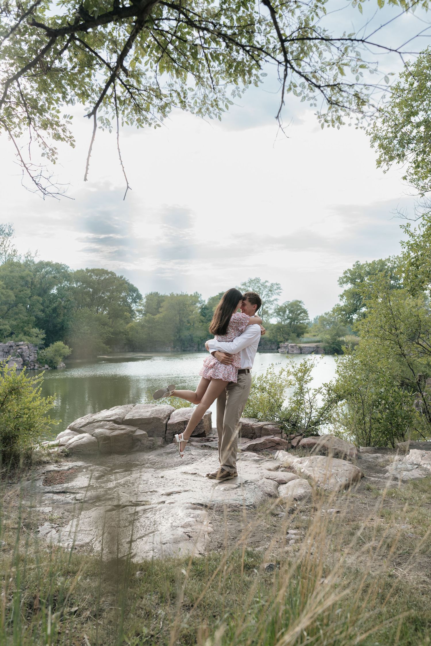 A couple shares a romantic embrace on rocky terrain beside a tranquil lake.