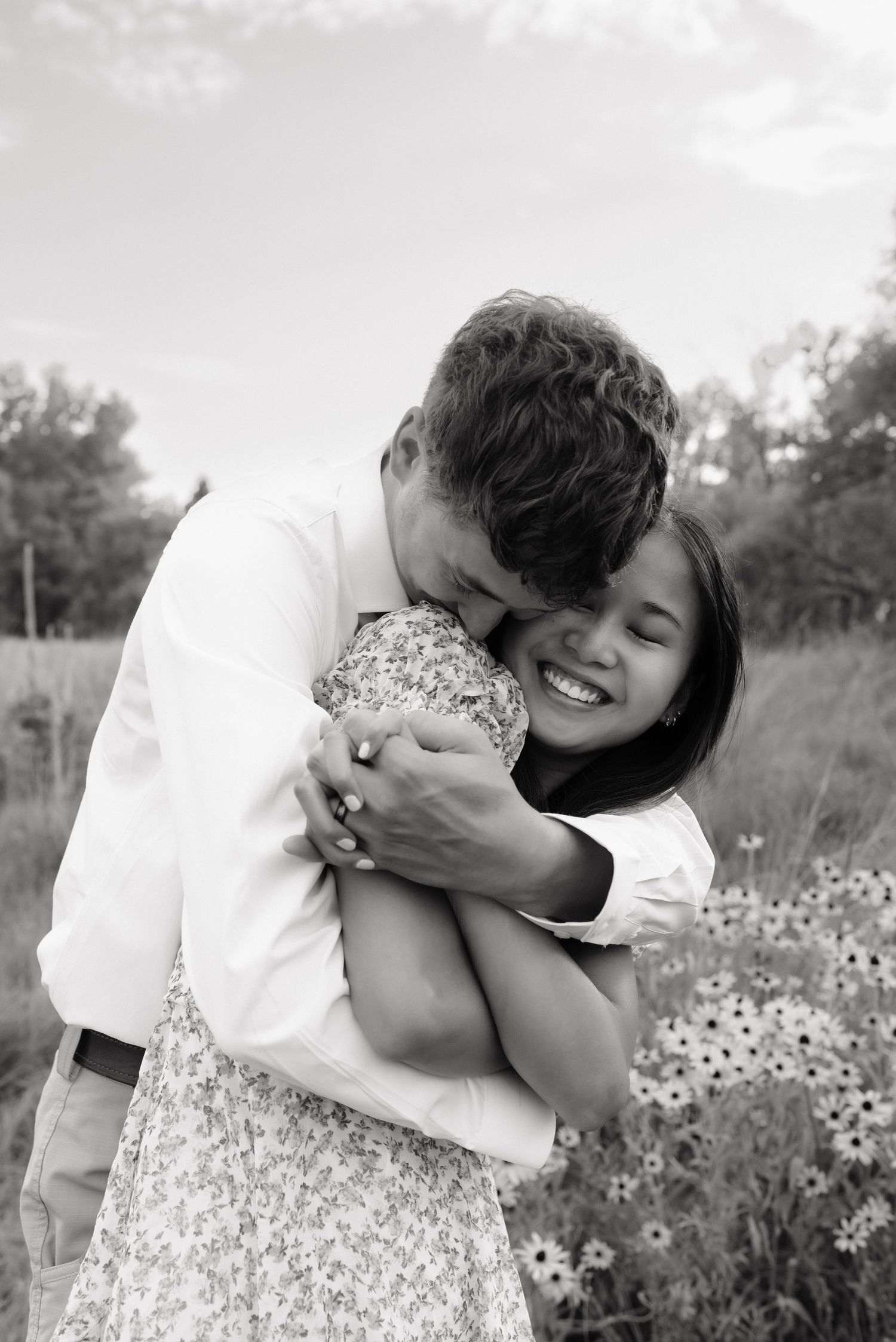 Black and white photo of a man hugging a laughing woman in a field of wildflowers.