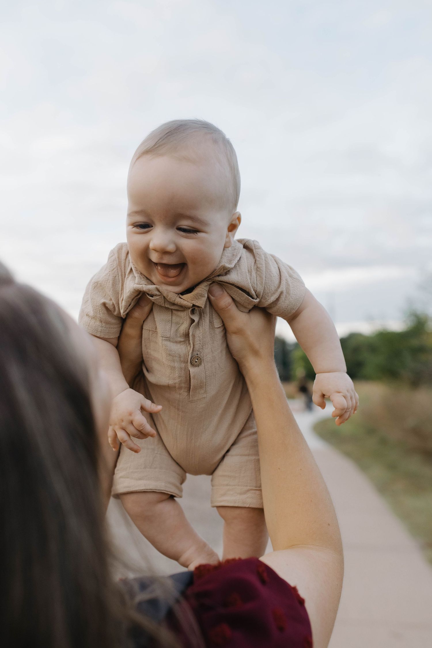 Joyful baby in beige romper laughing while being lifted up outdoors by a parent on a scenic path.
