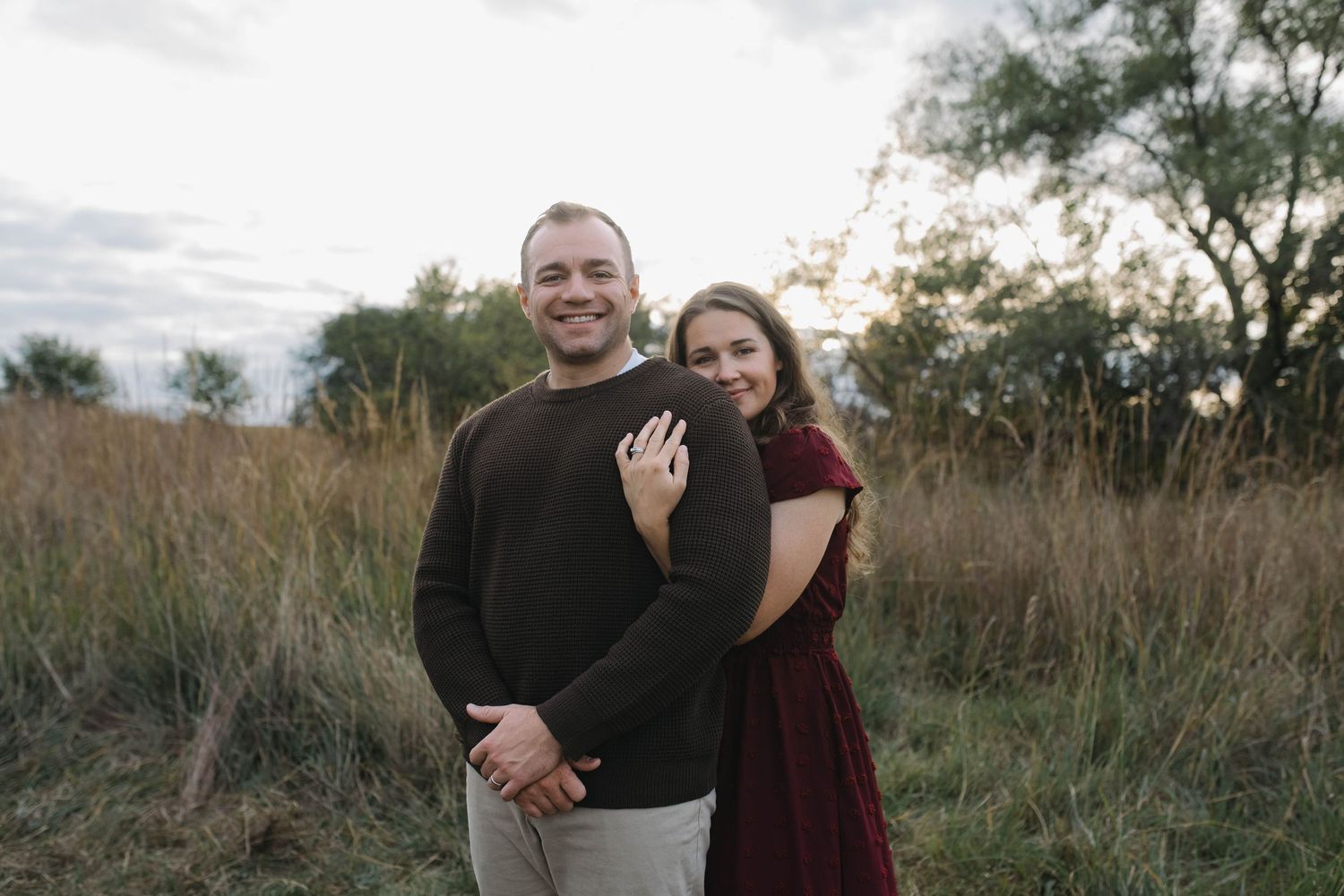 A woman in a burgundy dress hugs a man in a dark sweater while posing in an autumn field.