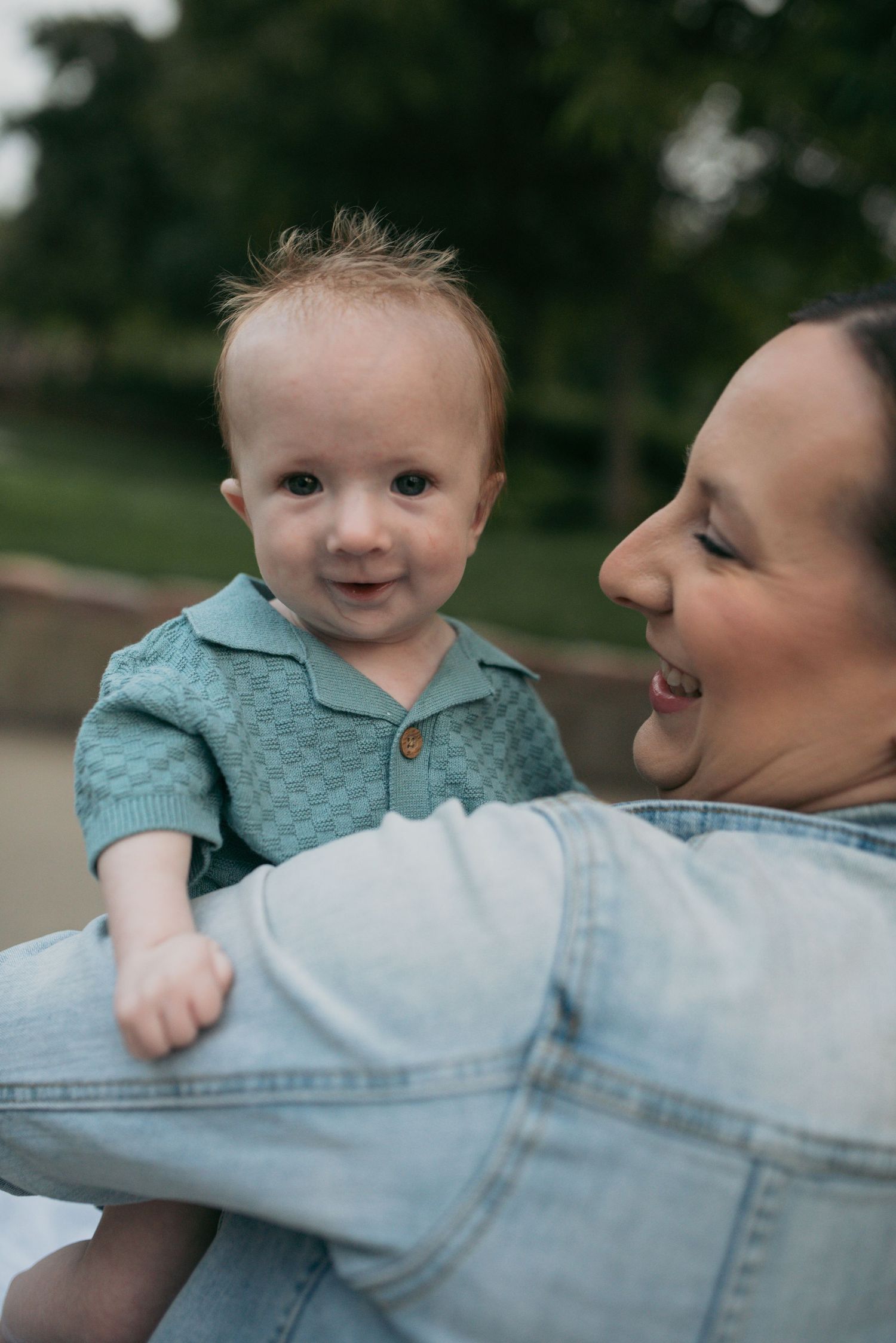 A smiling baby in a teal knit outfit being held by a laughing woman in a denim jacket.