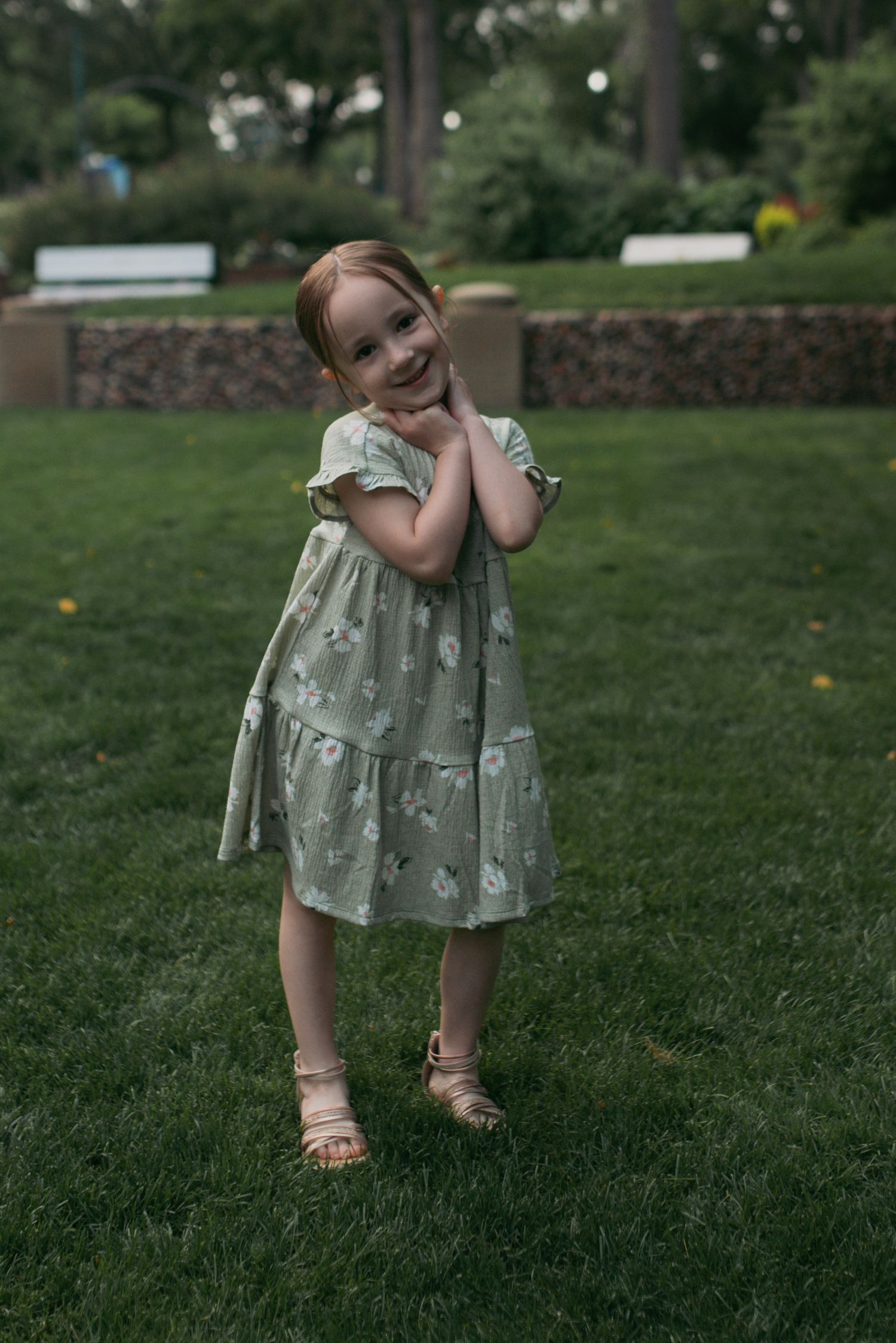 Cheerful young girl in mint floral dress posing with hands clasped on green lawn.