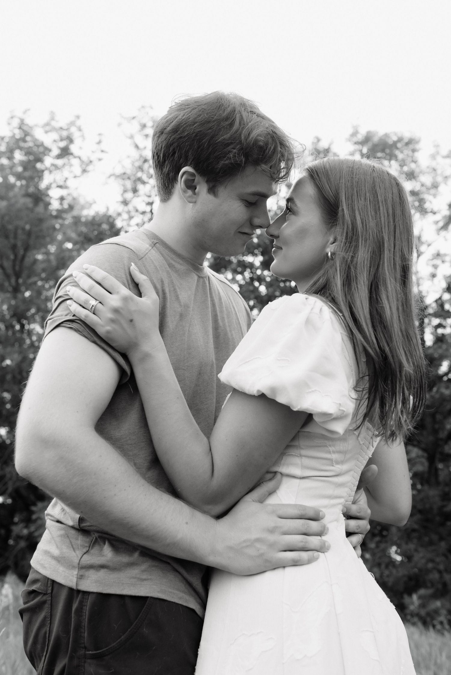 Black and white photo of a young couple embracing outdoors, about to kiss, surrounded by trees.