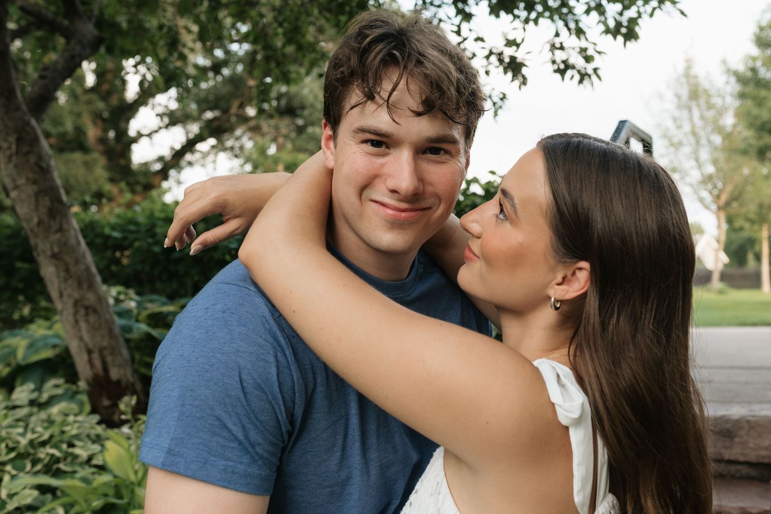 A young couple embracing outdoors, smiling warmly, with lush green trees in the background.