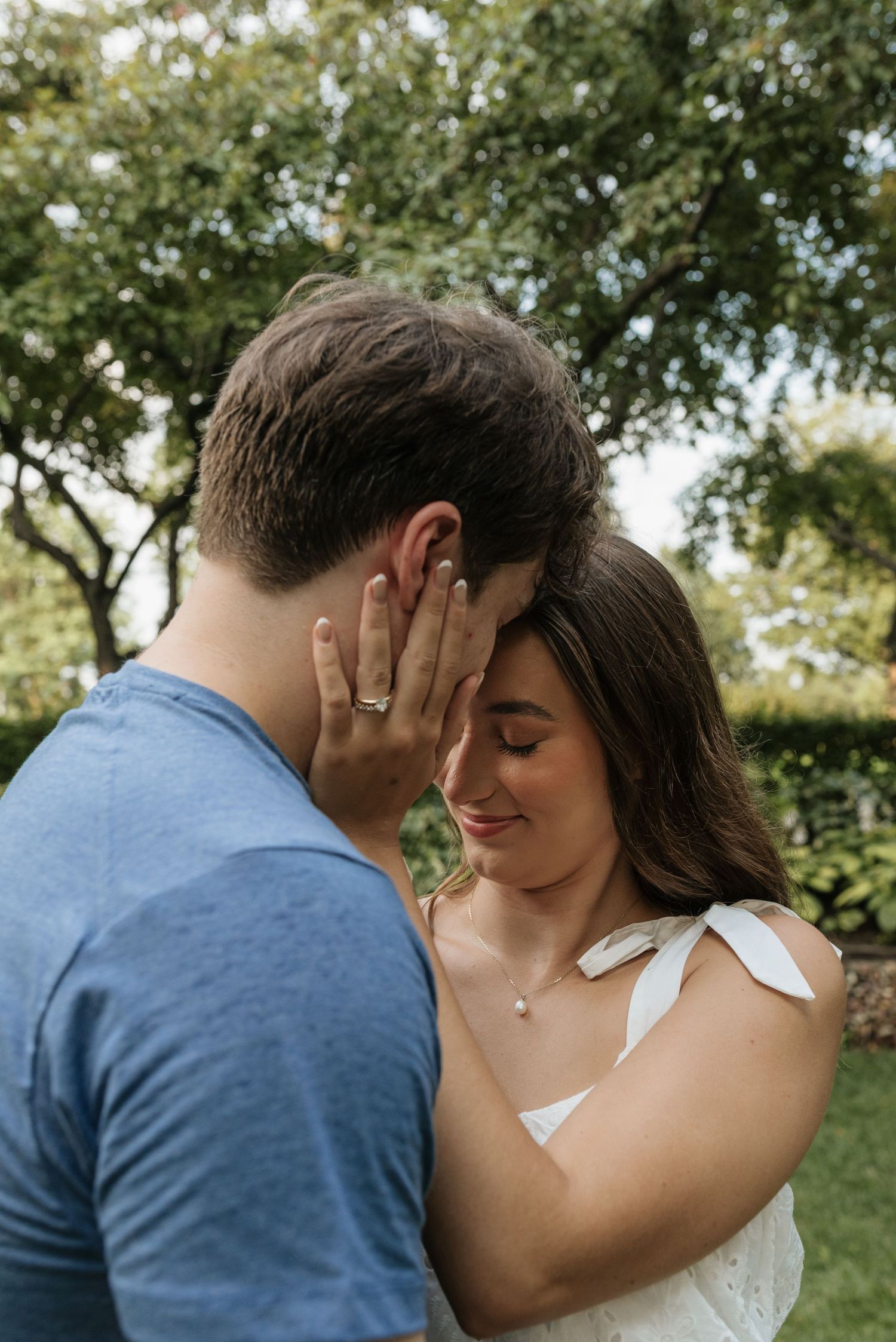 Couple sharing an intimate moment outdoors, woman smiling with engagement ring visible, lush green trees in background.