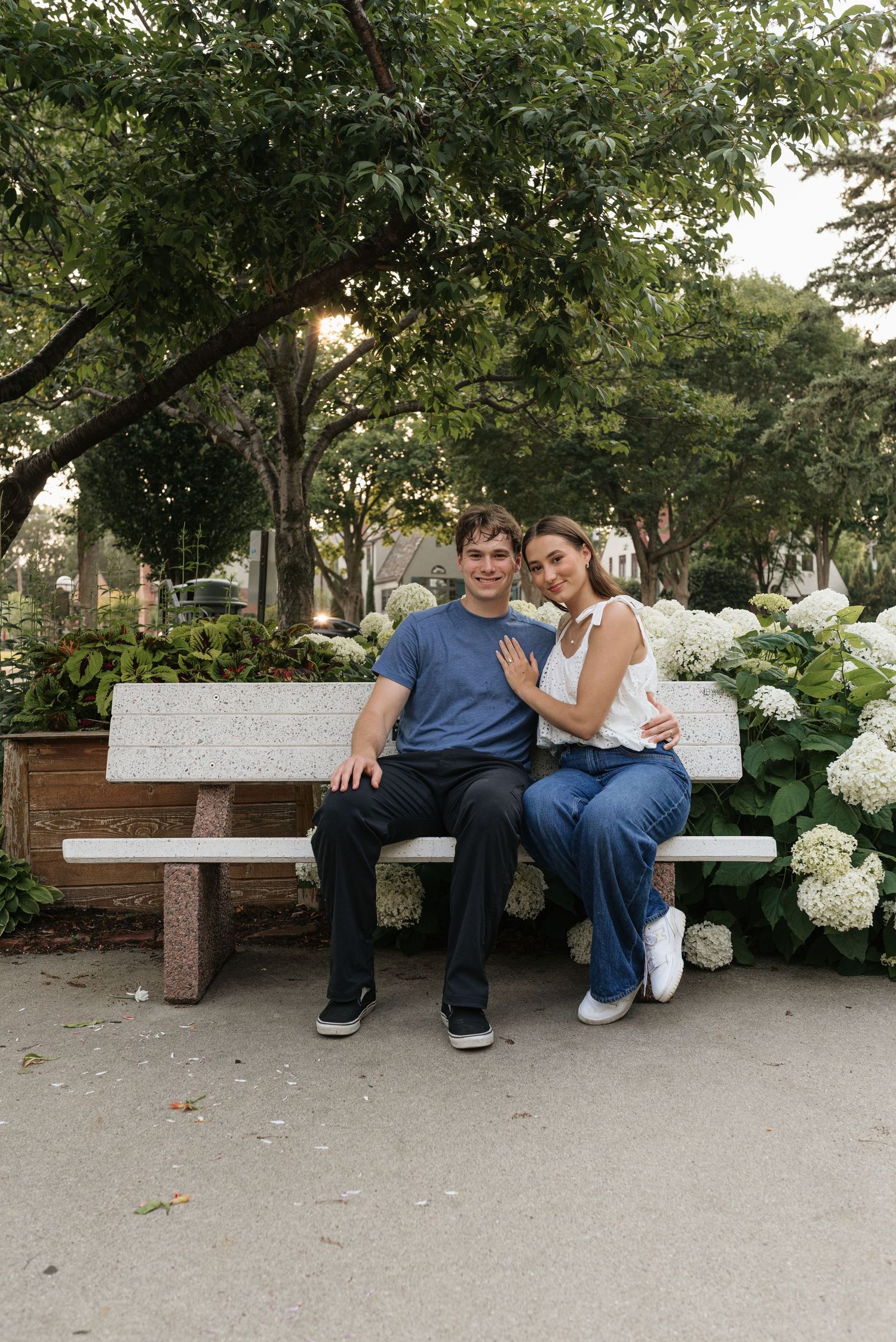 A couple sits closely on a white park bench surrounded by lush trees and white hydrangeas, smiling warmly at the camera.