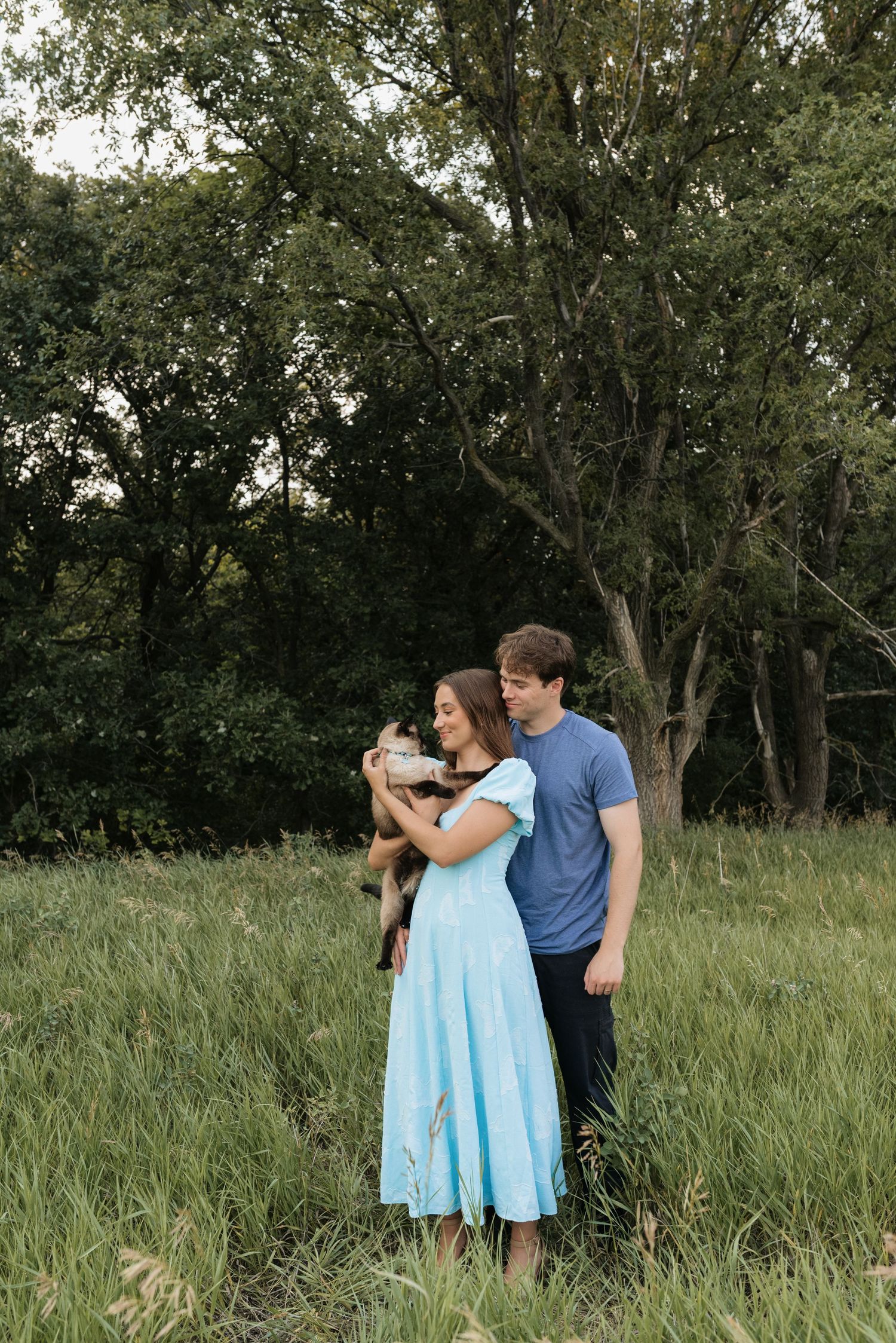 A couple stands in a green meadow holding a small dog, with lush trees in the background.