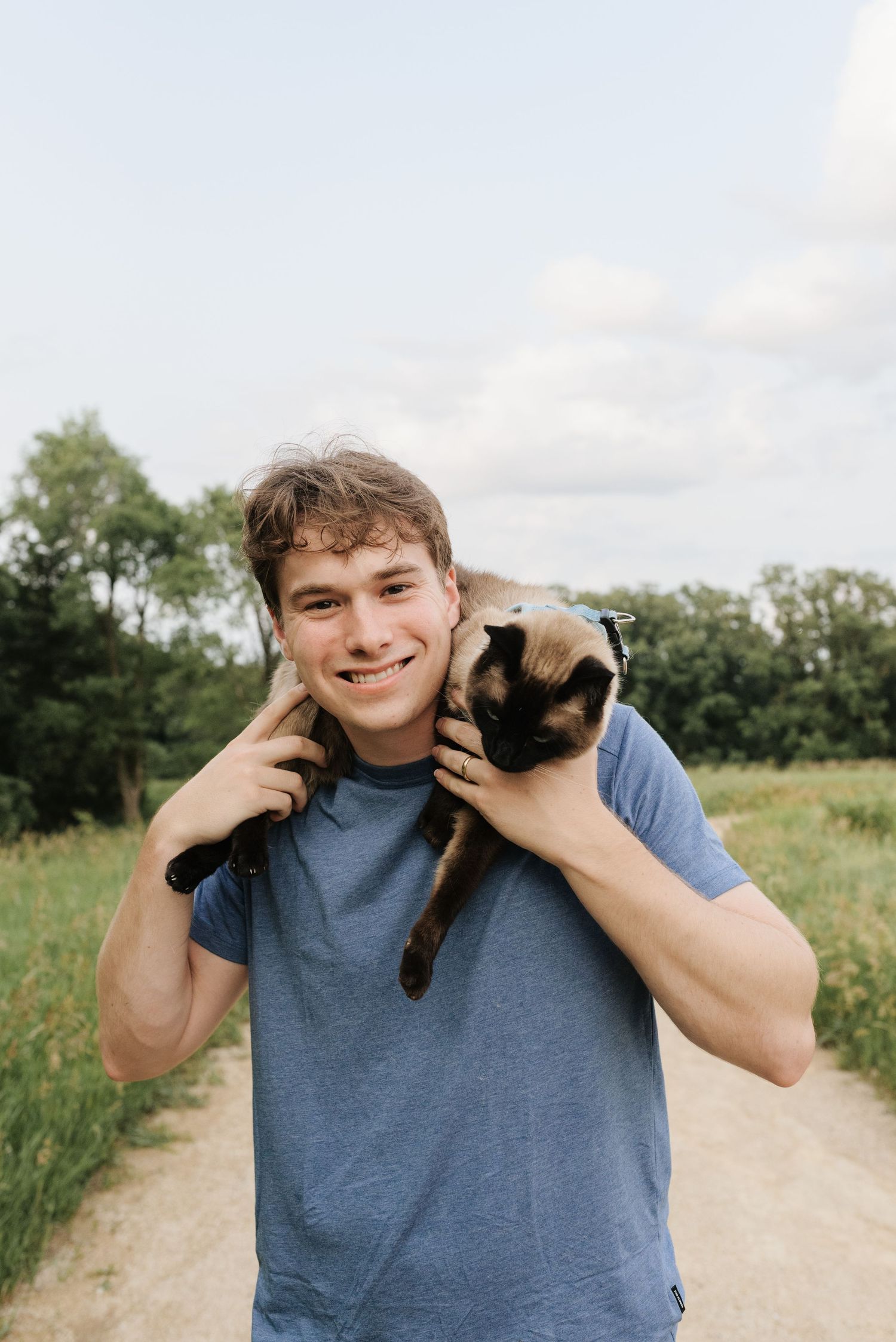 Young man smiling outdoors holding a Siamese cat on his shoulders on a rural path with green trees.