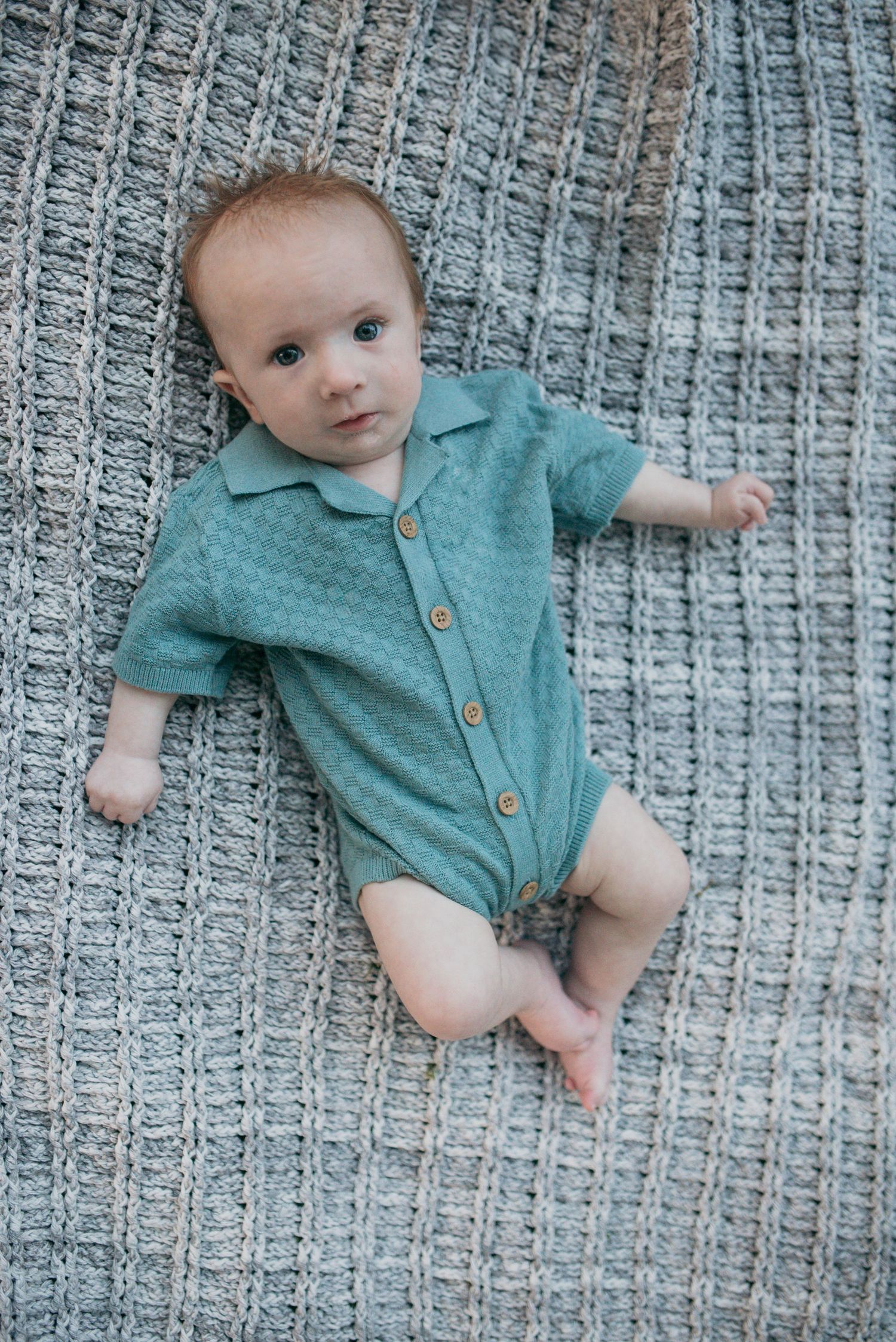 An adorable infant in a green collared onesie lying on a textured gray knit blanket.