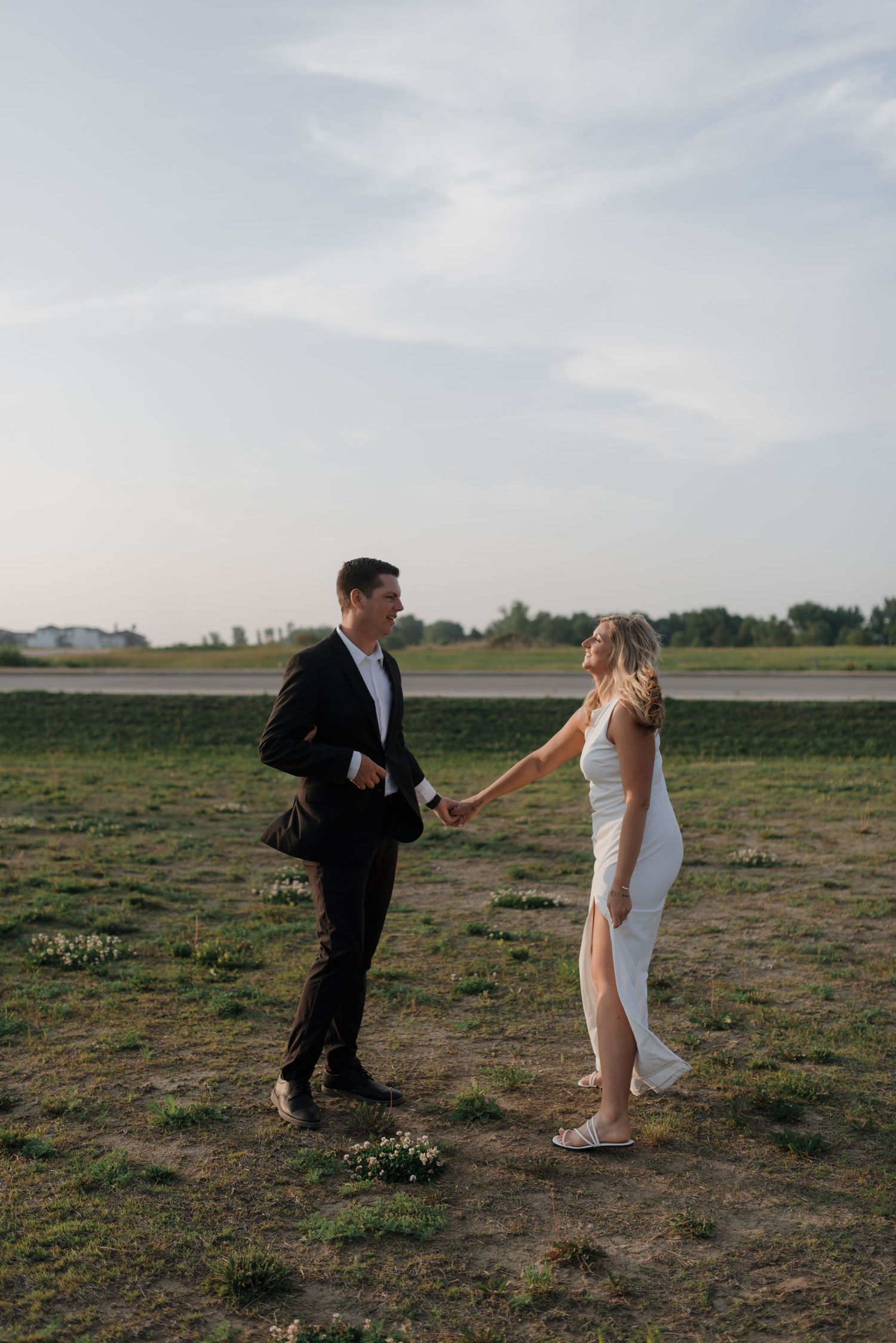 Couple holding hands and facing each other while smiling in open grassy field at sunset.