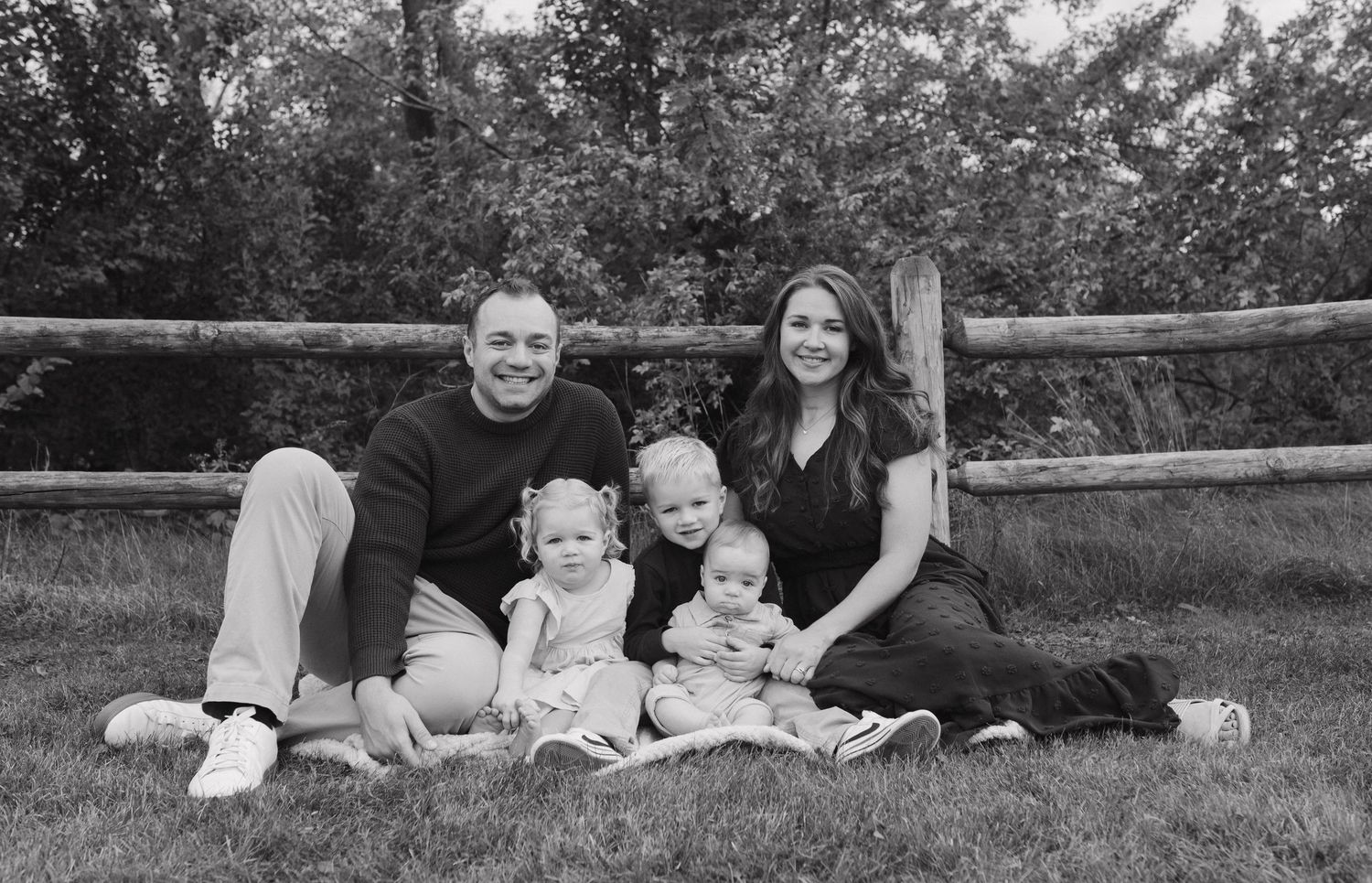 Happy family of five poses on green grass in front of a wooden fence in black and white.