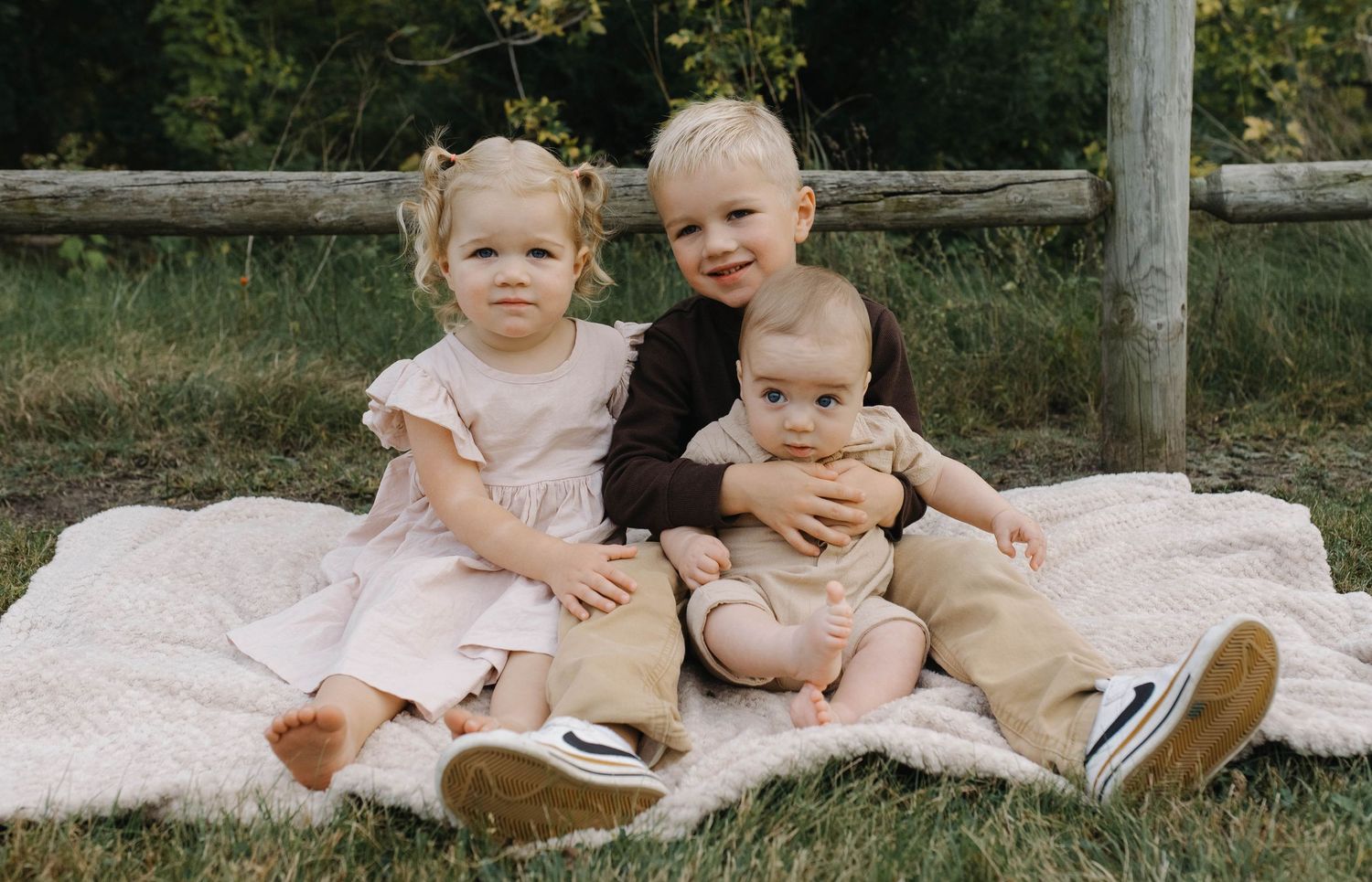 Three young children posing on a picnic blanket outside, with a wooden rail fence and green trees in the background.