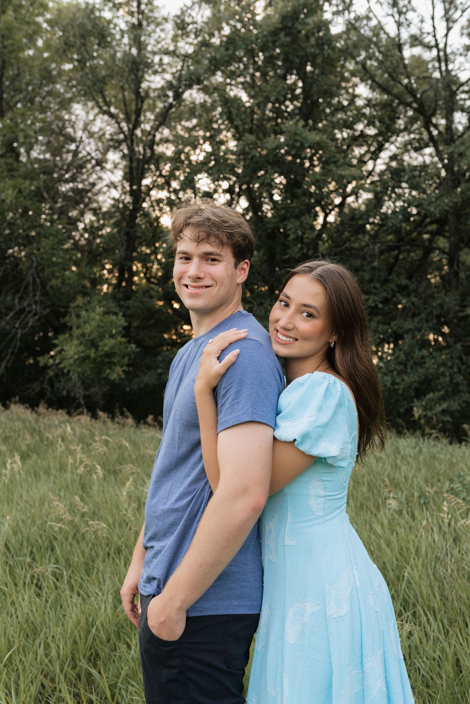 A smiling couple poses in a green meadow at sunset, woman in light blue dress embracing man in blue t-shirt.