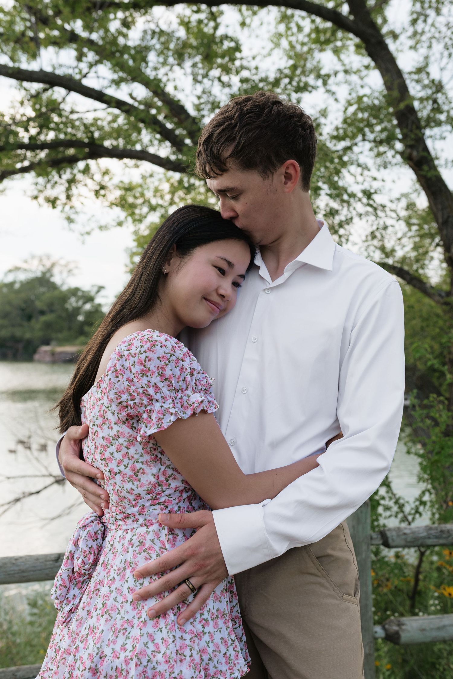 Couple embracing near a lake, woman in floral dress leans on man in white shirt among green trees.