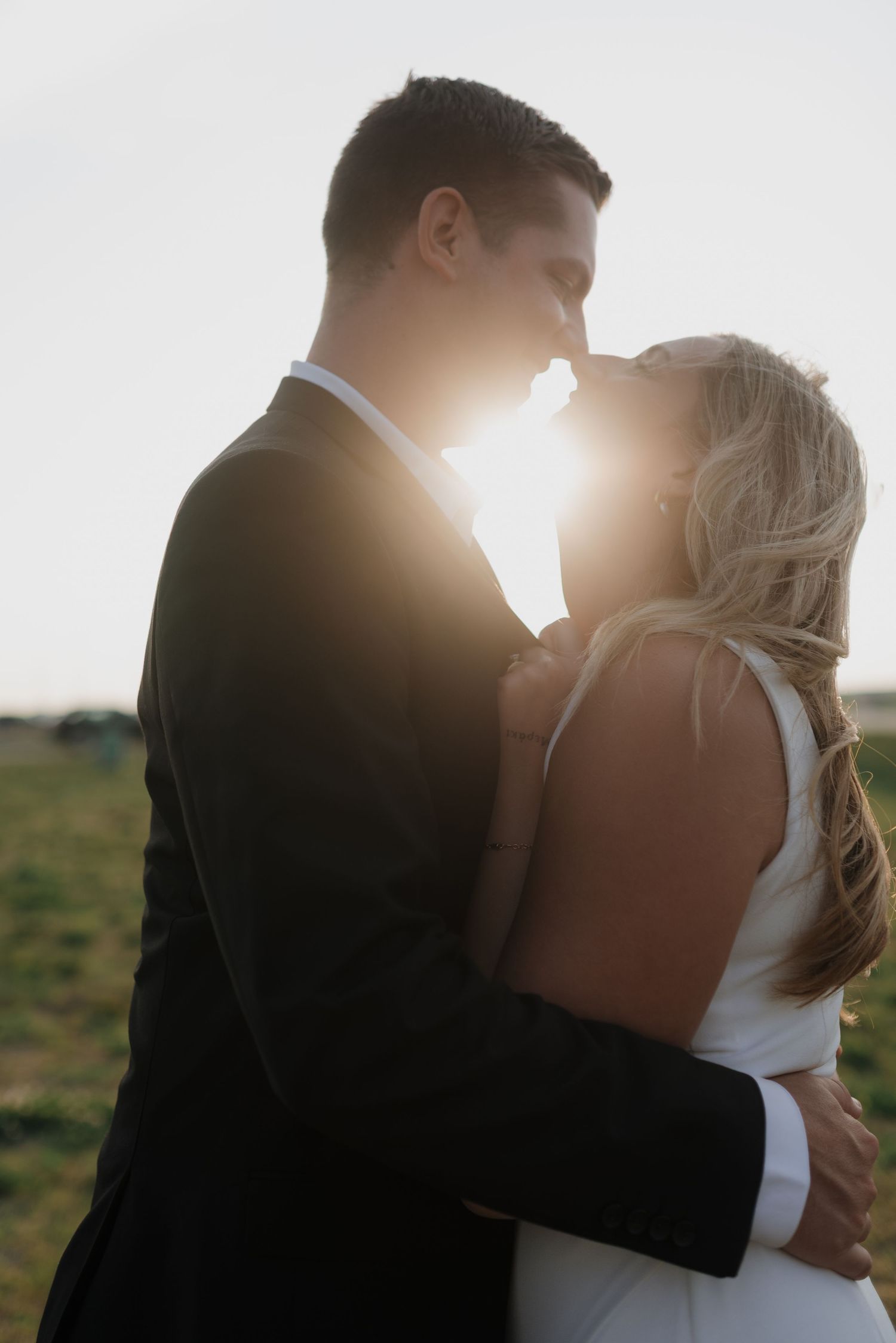 Bride and groom embracing face to face with radiant sunlight glowing behind them in an open field.