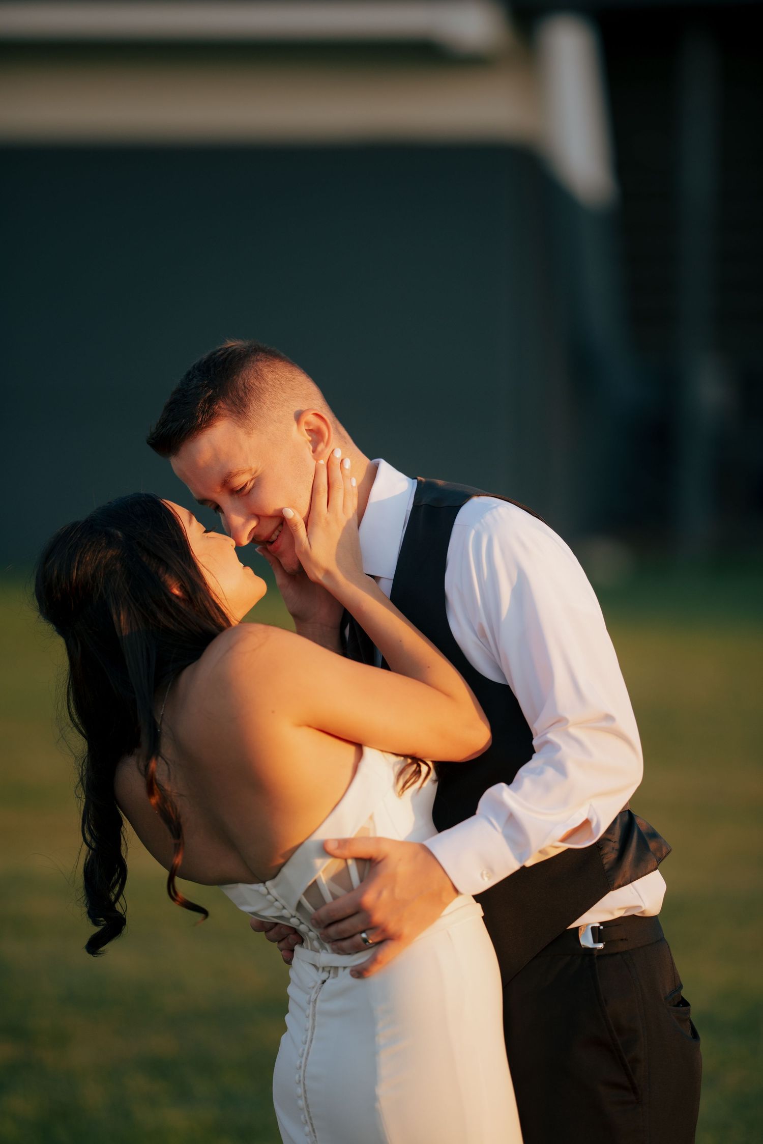 Romantic couple shares a kiss at golden hour, bride in white gown and groom in white shirt and vest.
