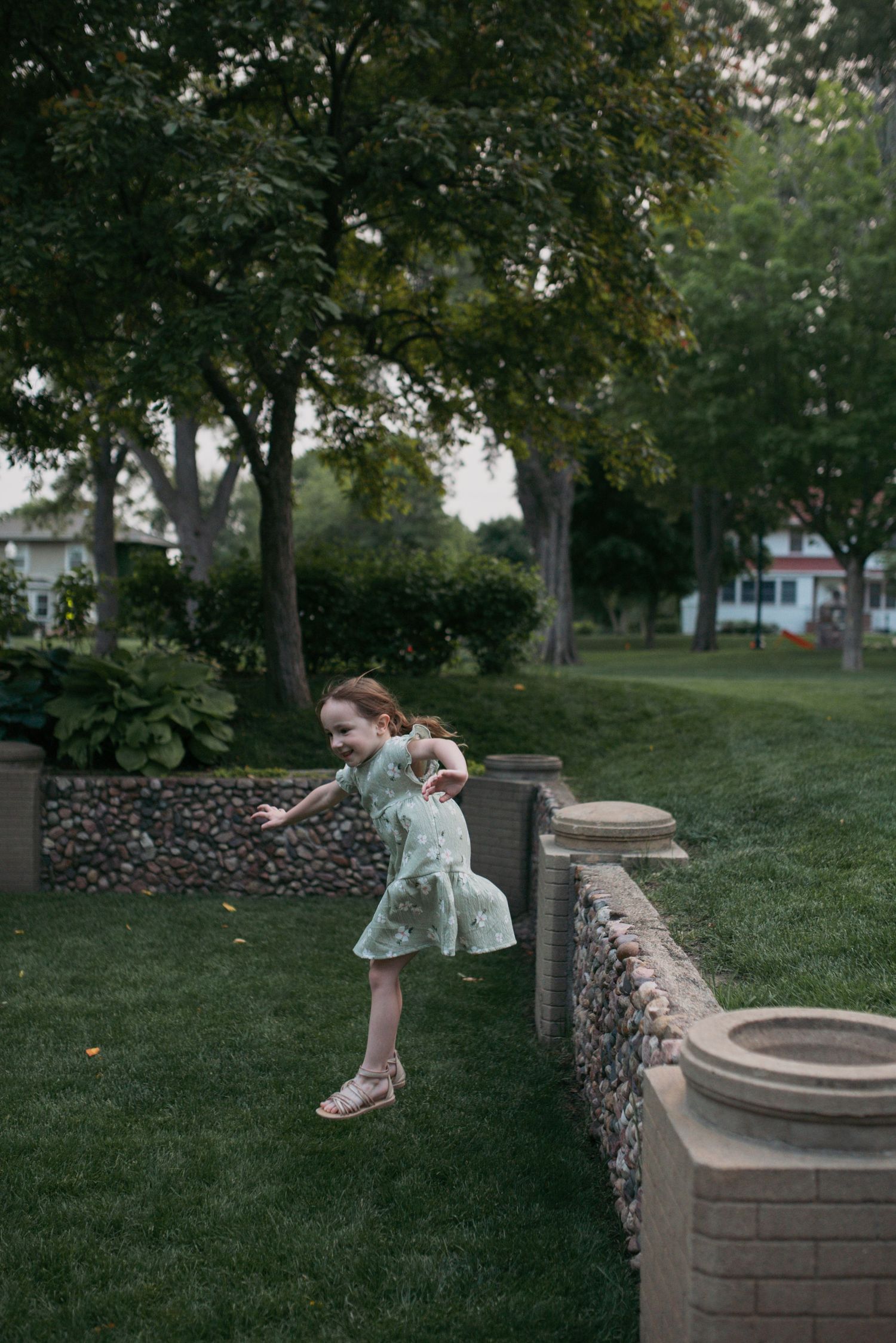 A young girl in a mint floral dress jumping off a stone garden wall in a lush green park.