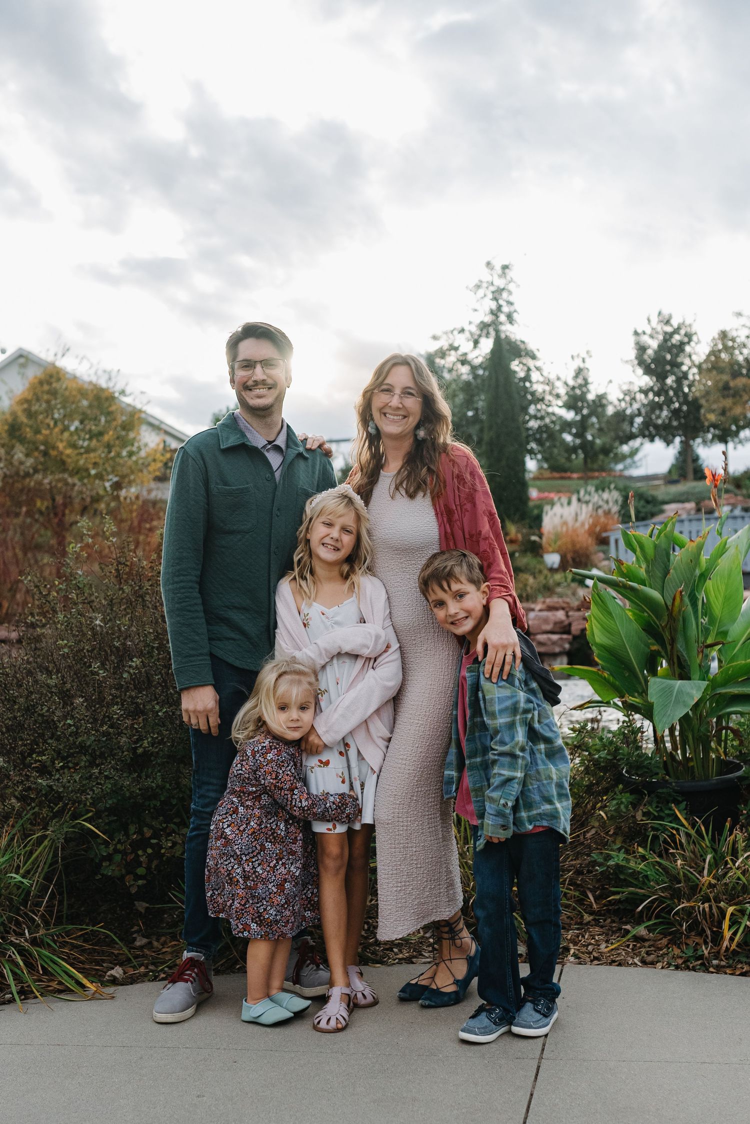 Family of five posing outdoors among colorful autumn garden plants on cloudy day.