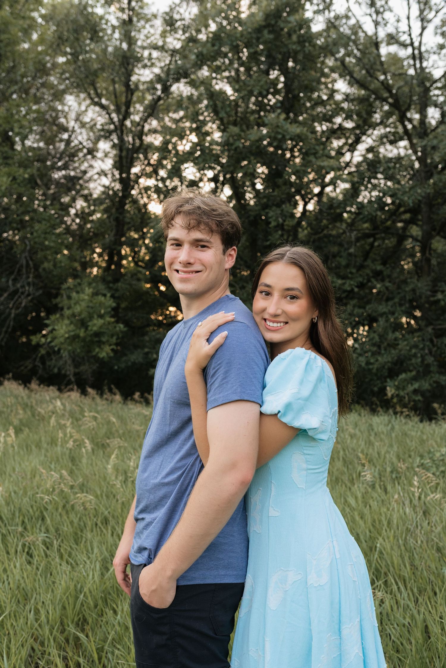 A young couple smiles outdoors in a green meadow, she wears a light blue dress, he wears a blue t-shirt.