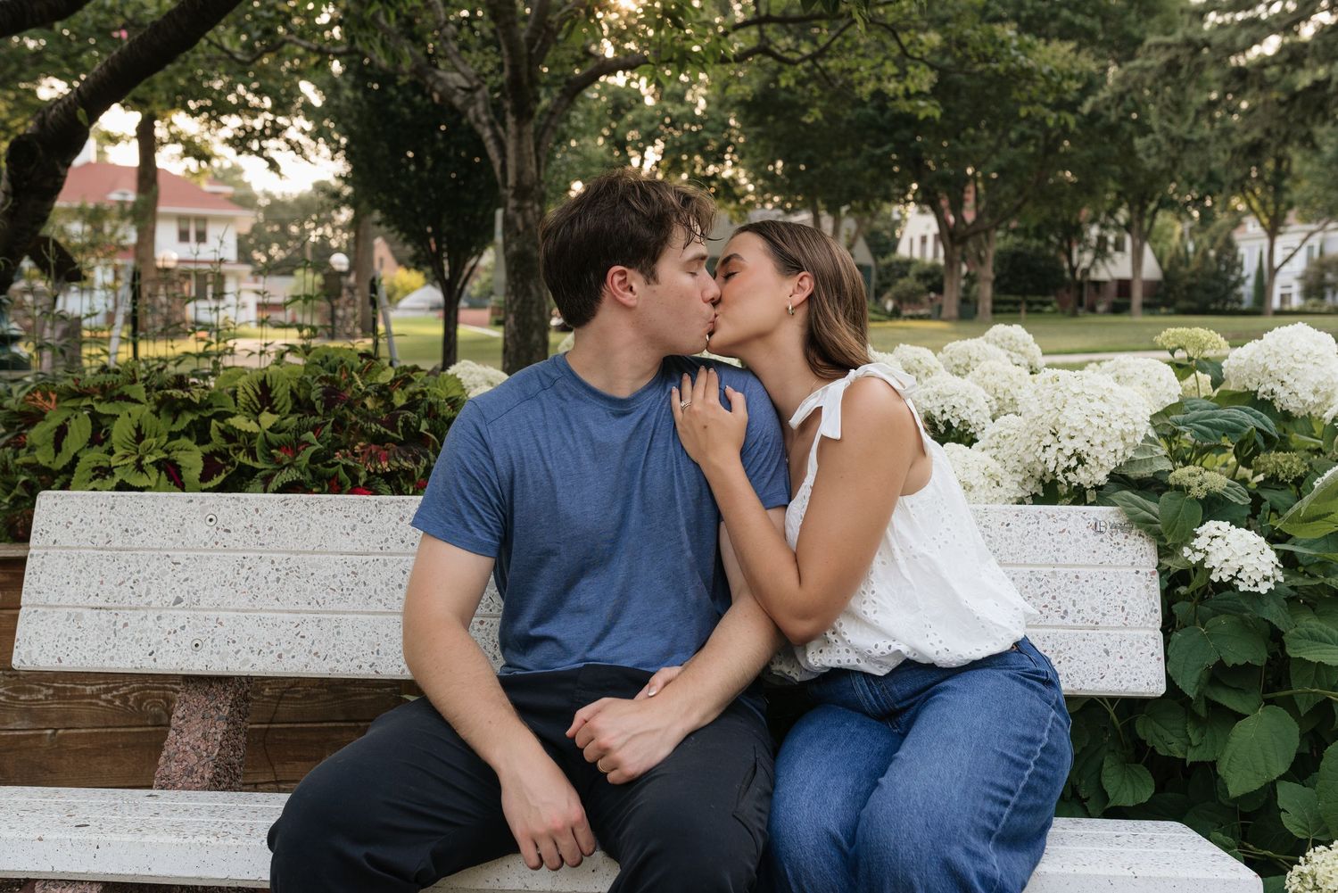 A couple shares a romantic kiss on a white park bench surrounded by white hydrangeas and lush greenery.