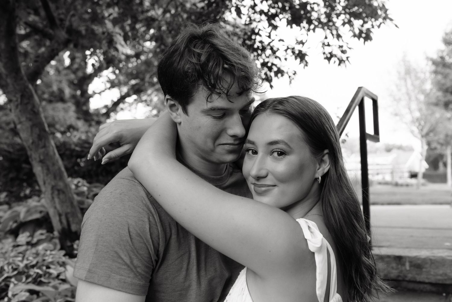 Black and white photo of a young couple embracing outdoors, woman smiling and hugging man near trees and steps.