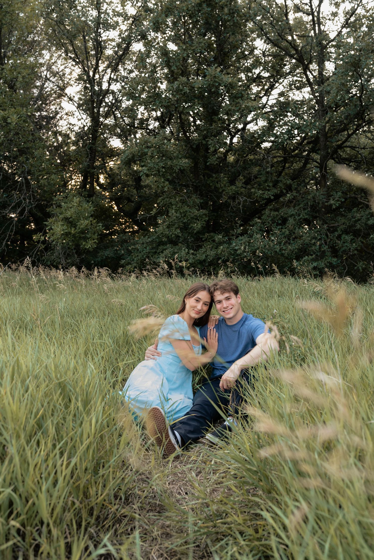 A couple sitting together in a lush green meadow, smiling at the camera, surrounded by tall grass and trees.