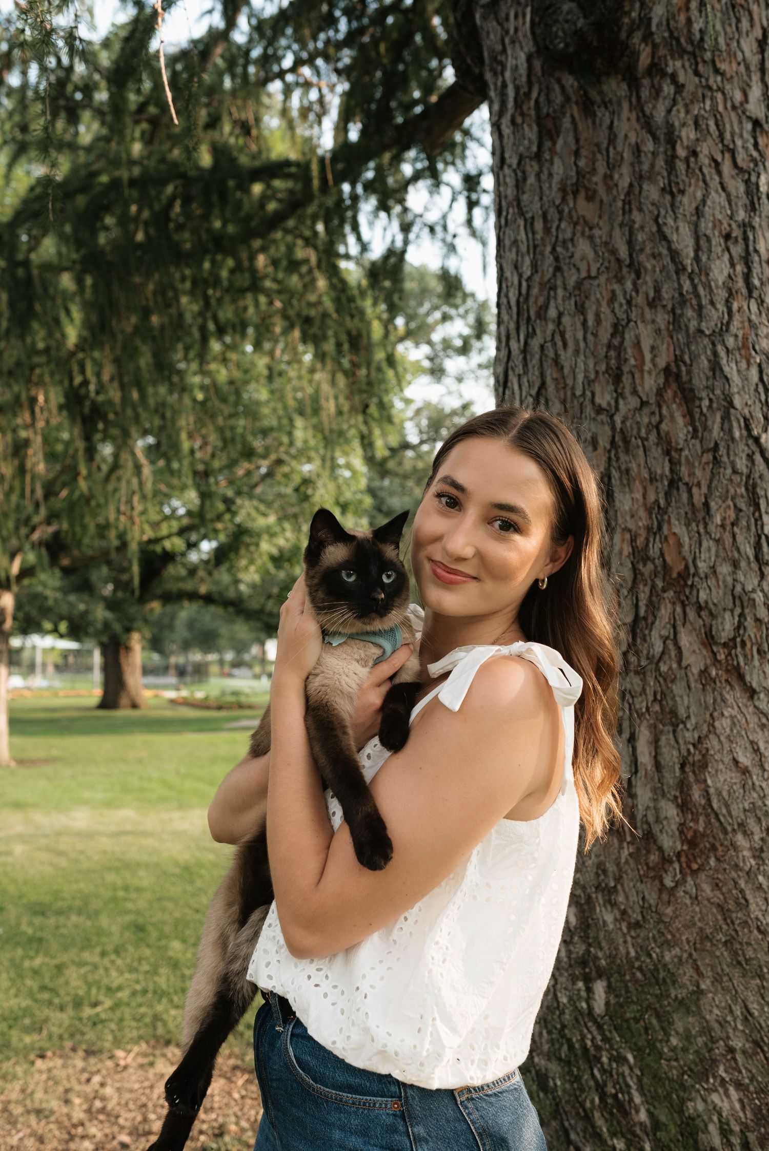 Young woman in white top holds a Siamese cat outdoors near a large tree in a lush green park.
