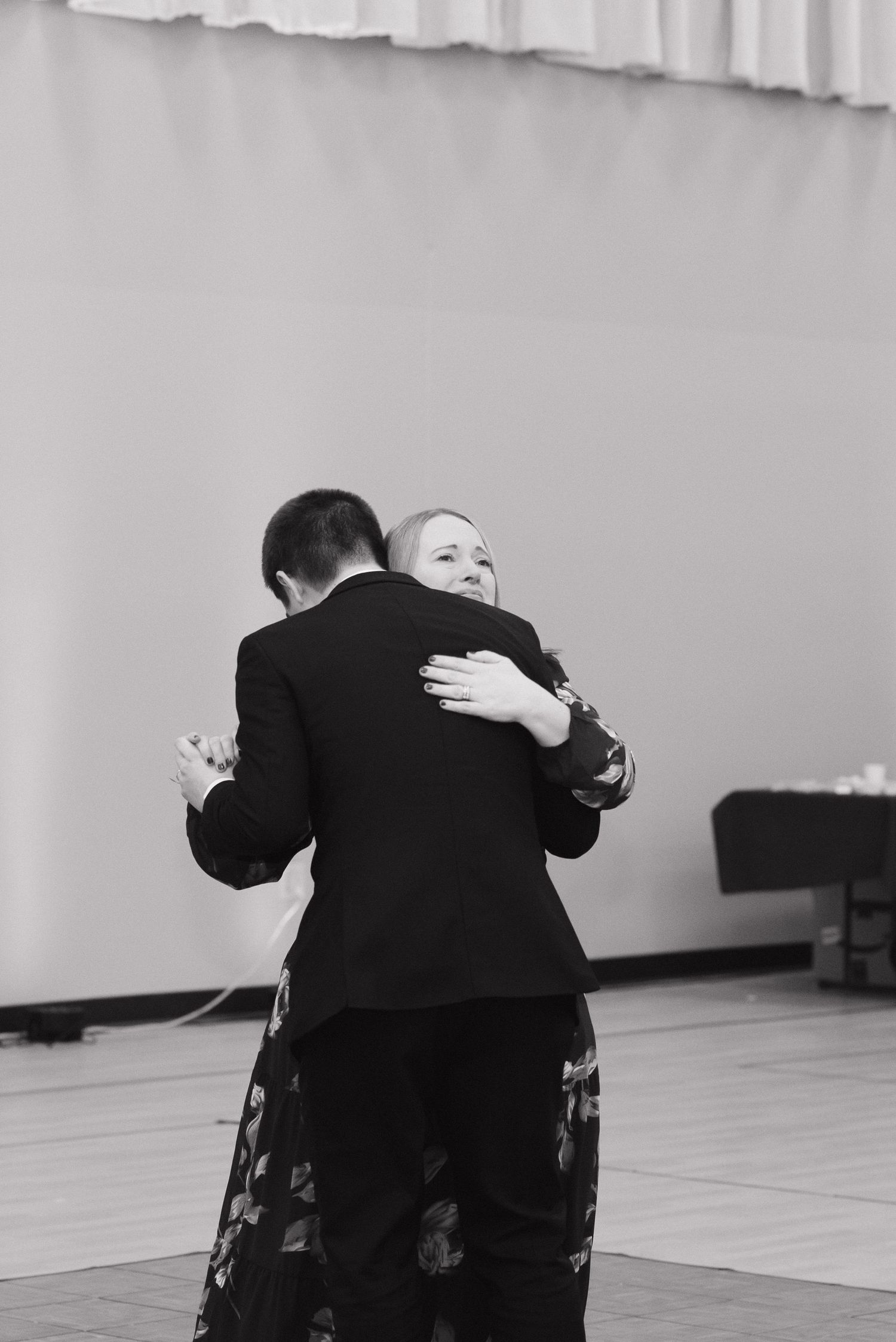 Groom and older woman share emotional dance embrace at wedding reception in black and white.