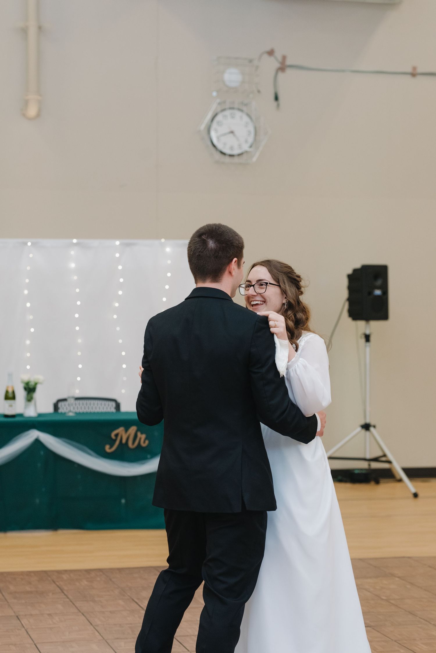 Bride in white gown and groom in black suit share first dance at wedding reception with fairy lights backdrop.