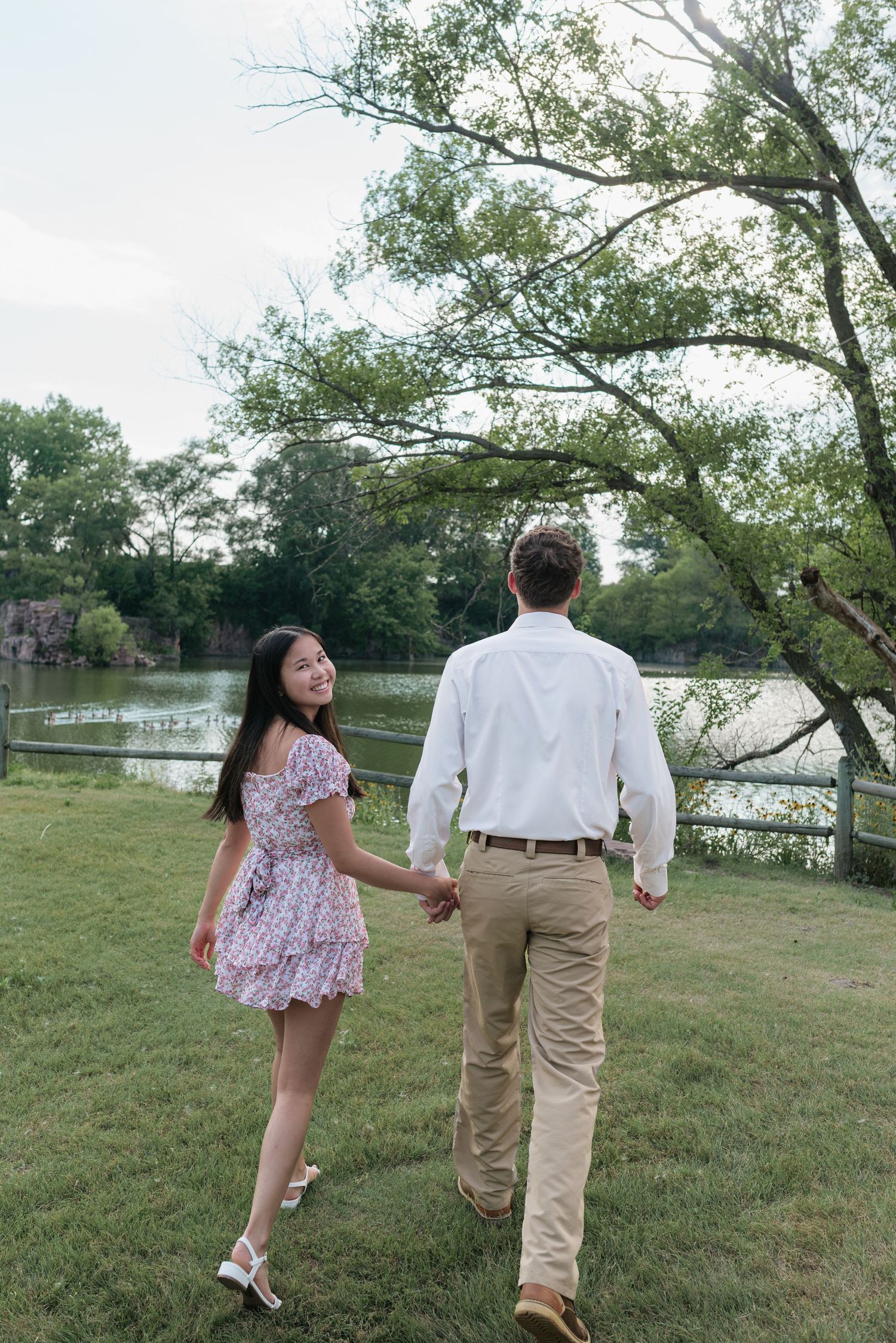 A smiling woman in a floral dress holds hands with a man in a white shirt near a scenic pond surrounded by trees.