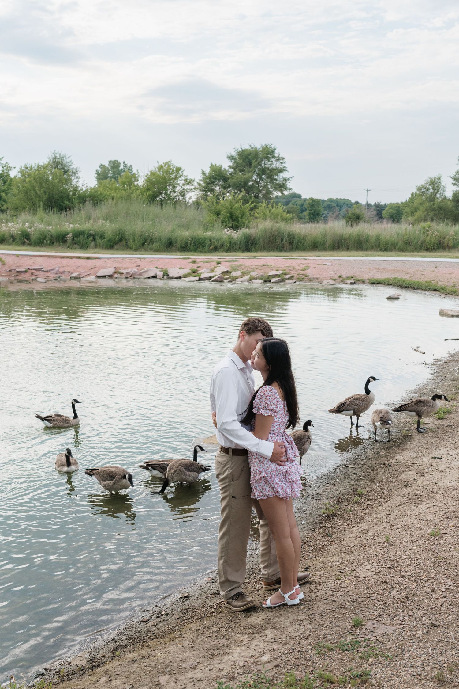 A couple shares a kiss by a peaceful pond with Canada geese along the shoreline.