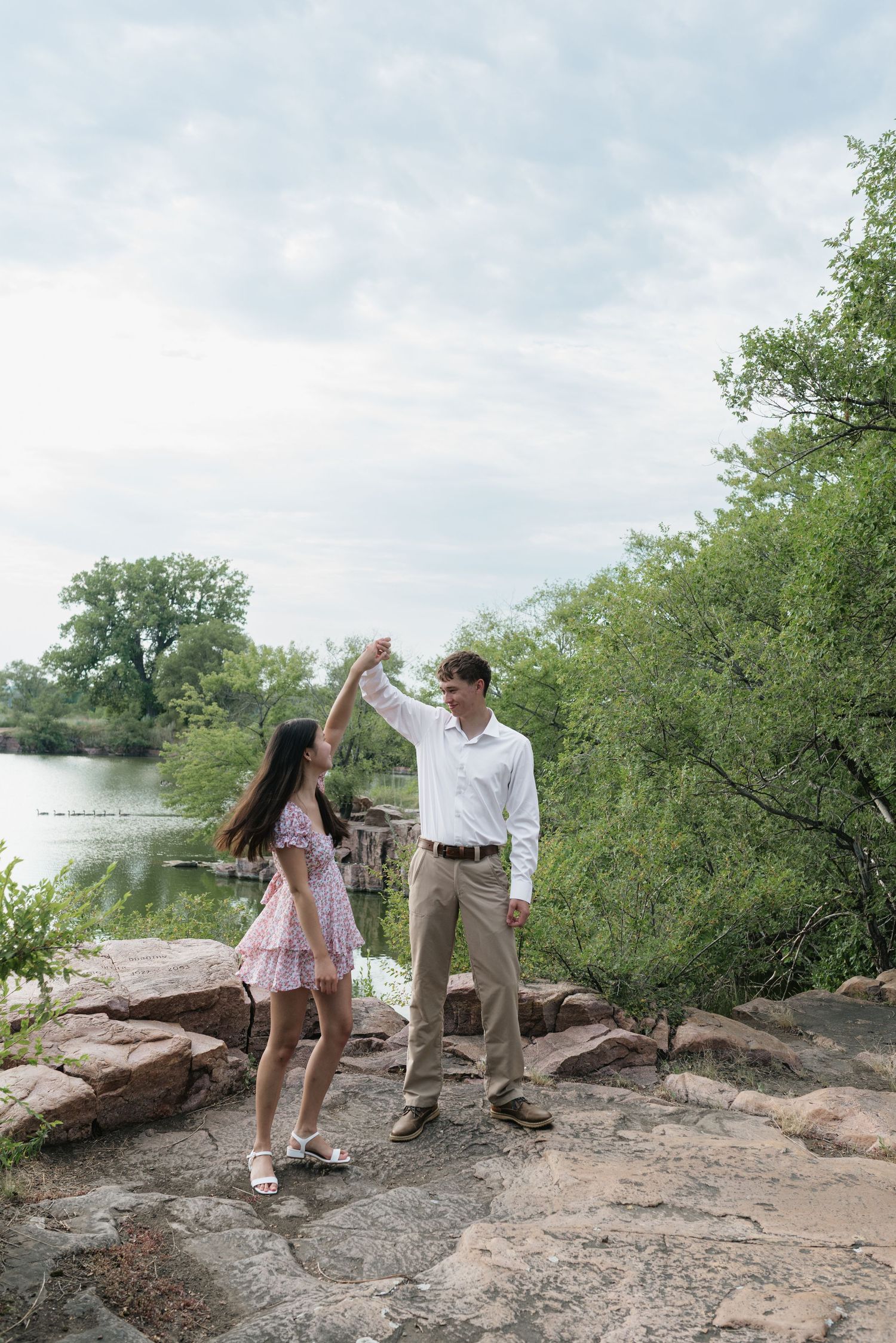Couple dancing on rocky lakeside with lush green trees and cloudy sky backdrop.