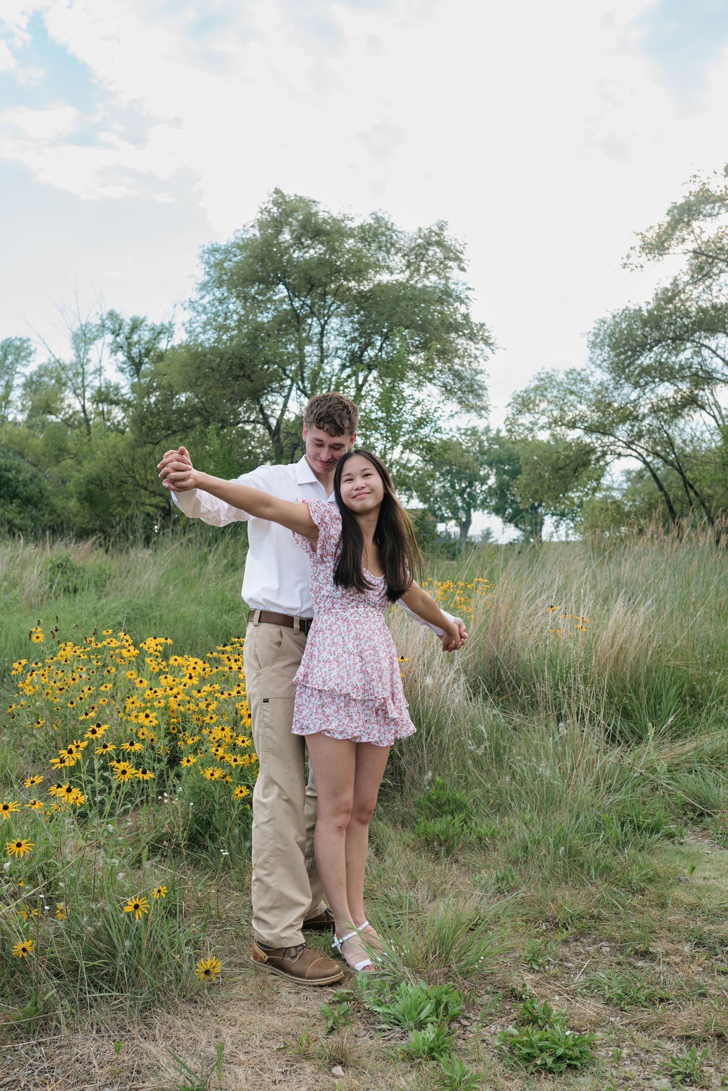 Couple dancing joyfully in a meadow with yellow wildflowers and lush green trees.