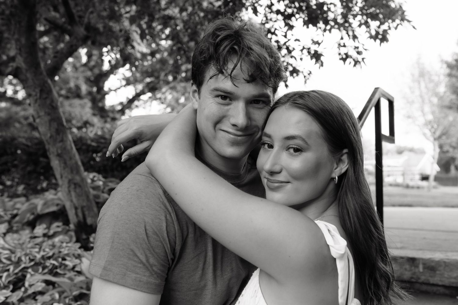 Black and white photo of a young couple smiling and embracing outdoors near trees and steps.