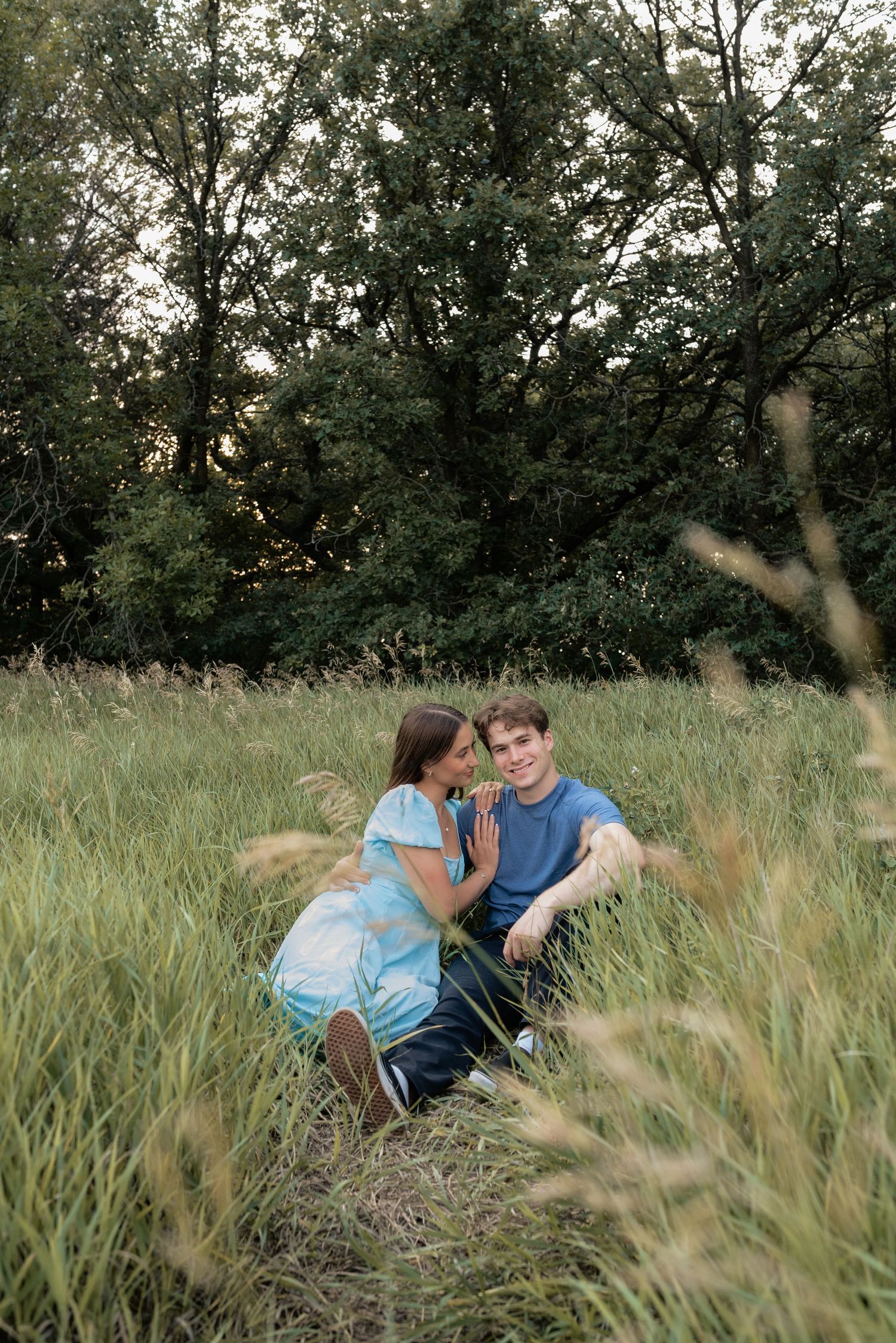 A couple sits together in a lush green meadow, sharing a tender moment surrounded by tall grass and trees.