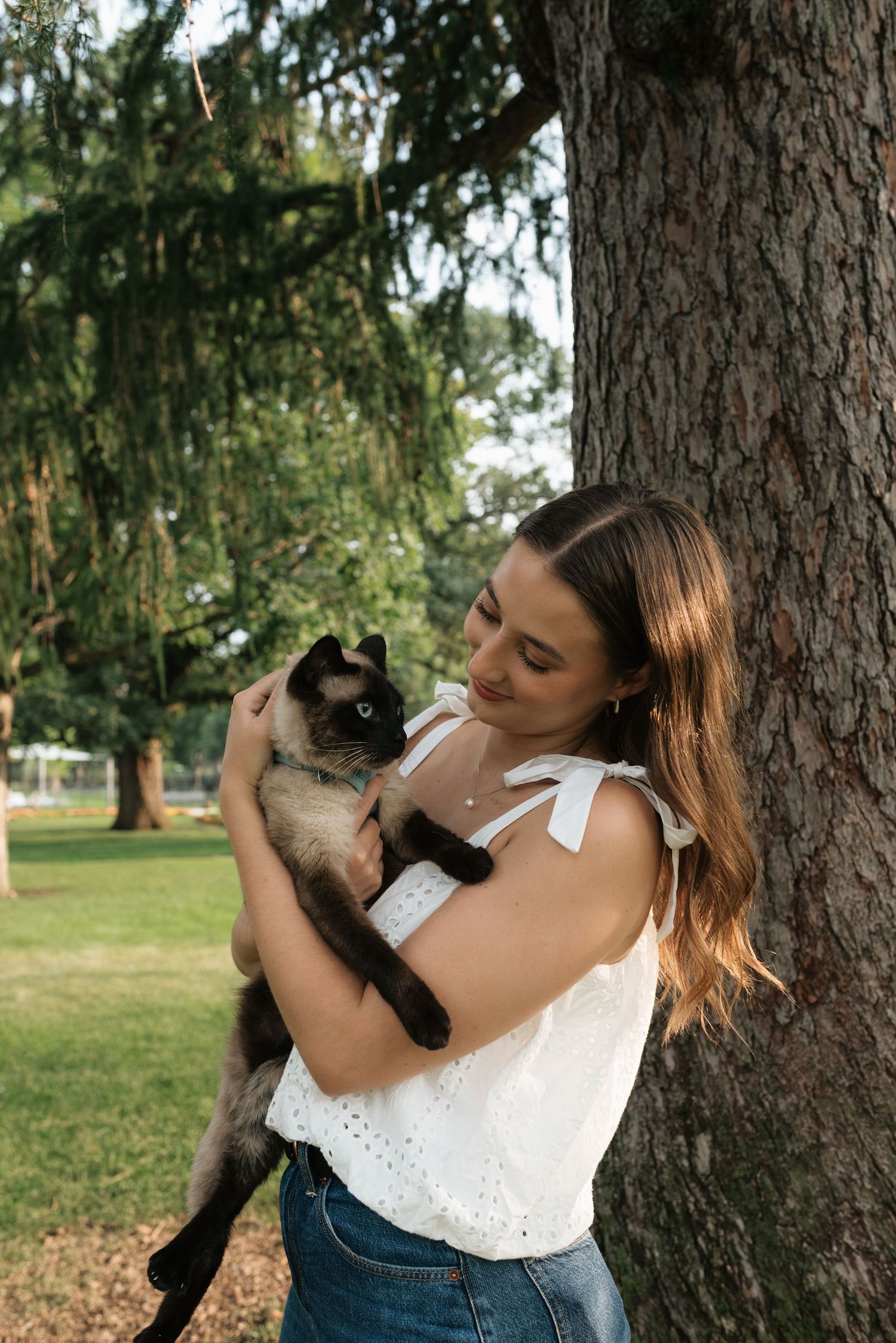 Young woman in white top holding a Siamese cat while leaning against a tree in a lush green park.