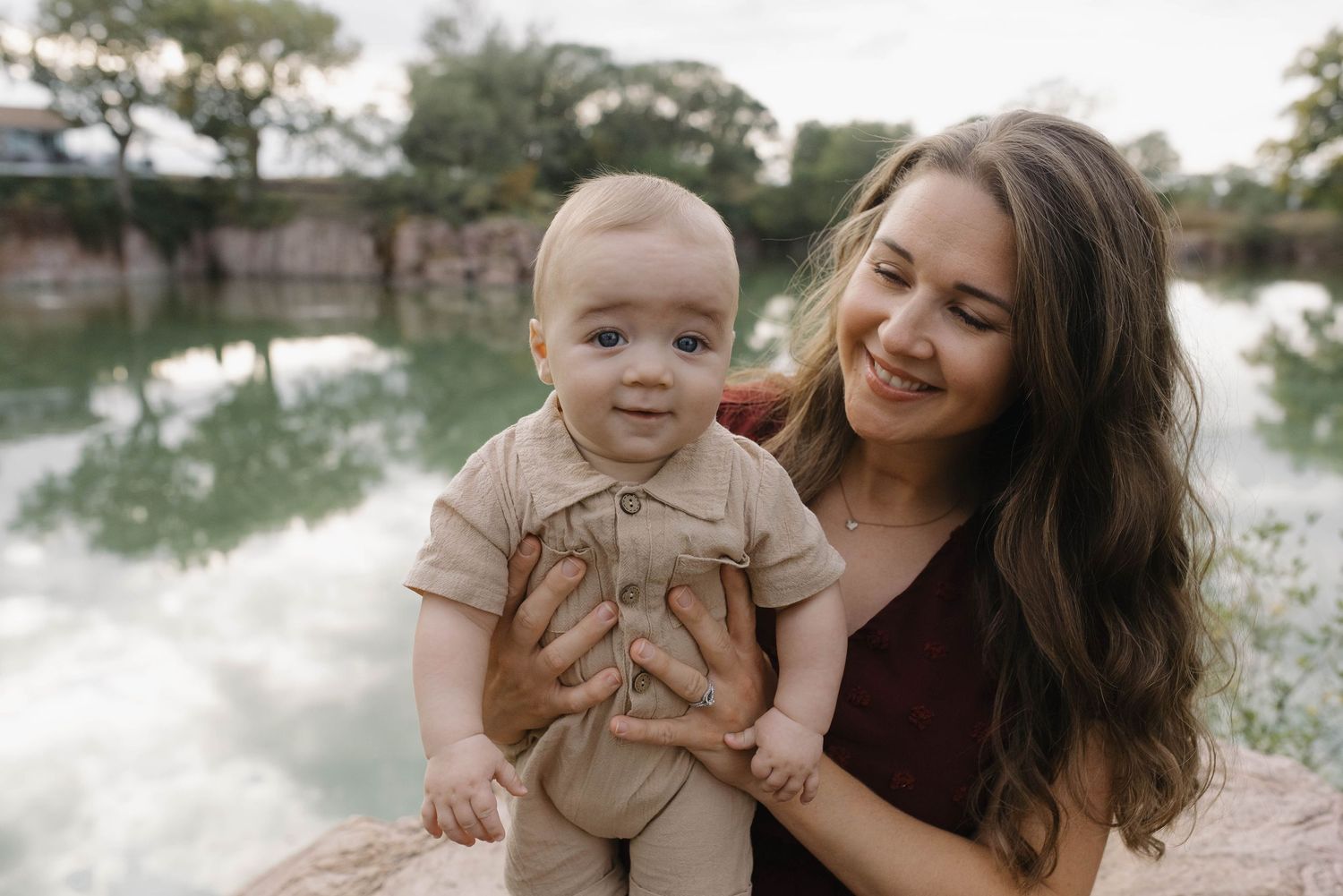 Smiling mother in burgundy dress holding an adorable baby boy near a reflective pond outdoors.
