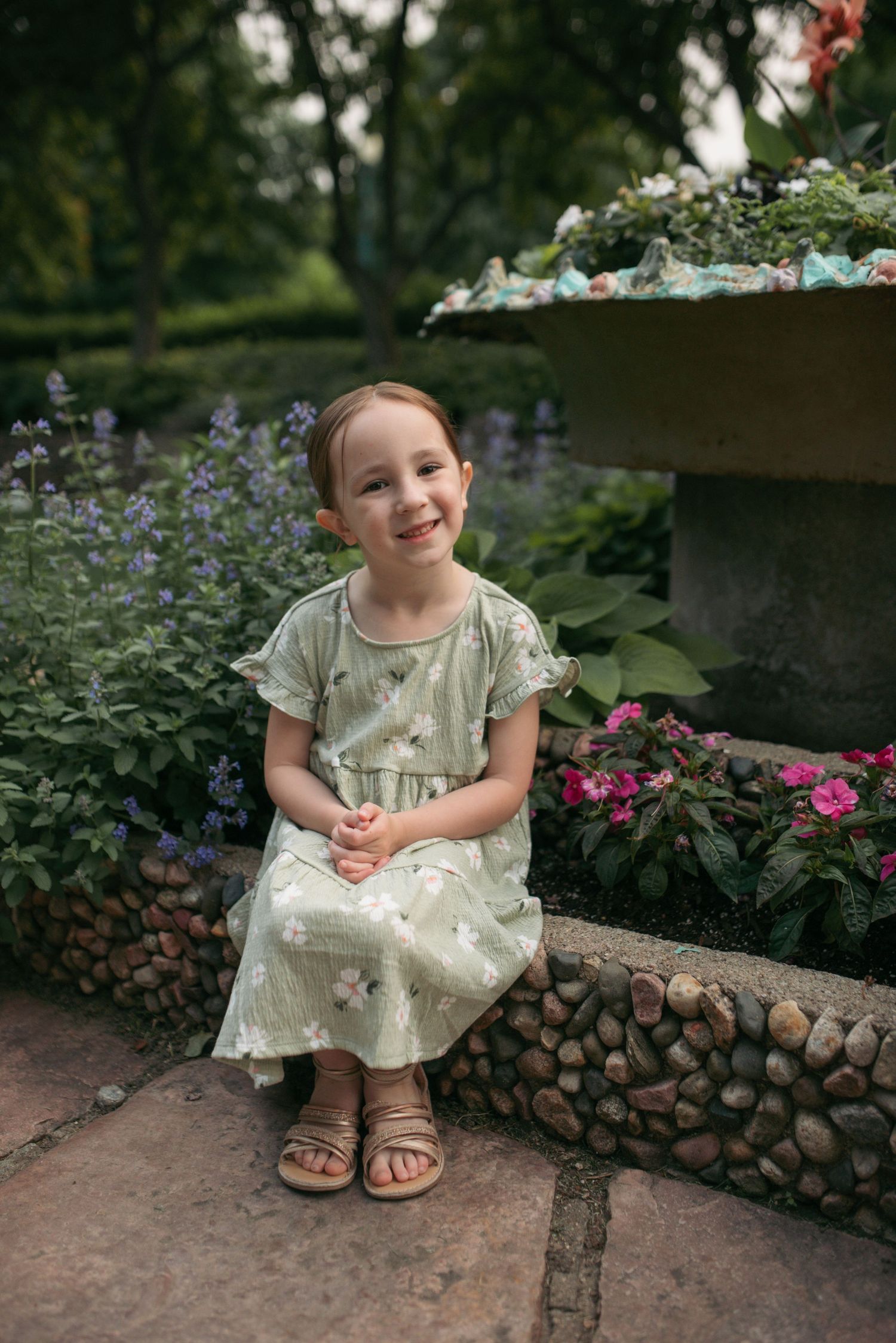 Happy young girl in light green floral dress sits in lush garden setting.