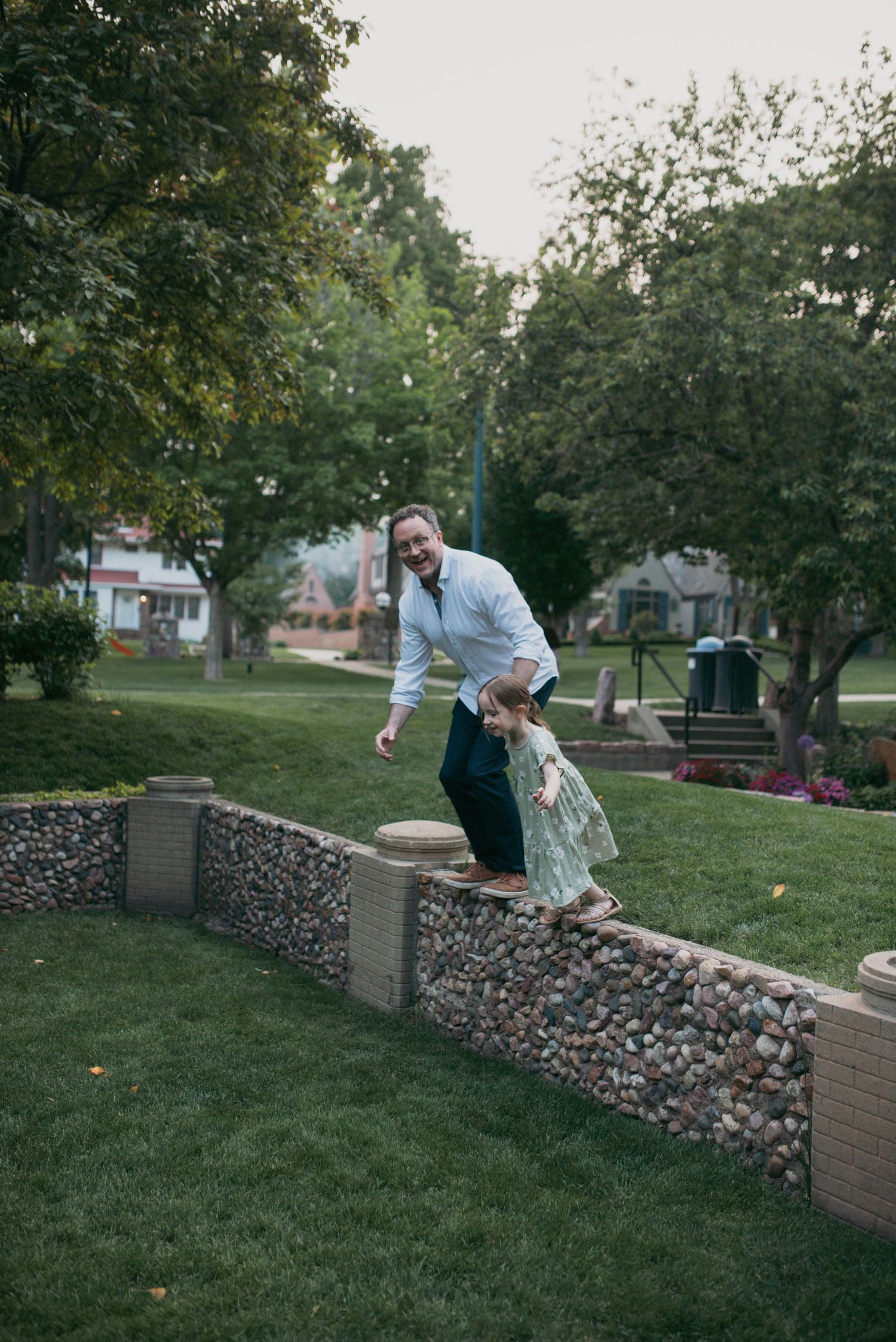 A father helping his young daughter walk along a stone garden wall in a green park.
