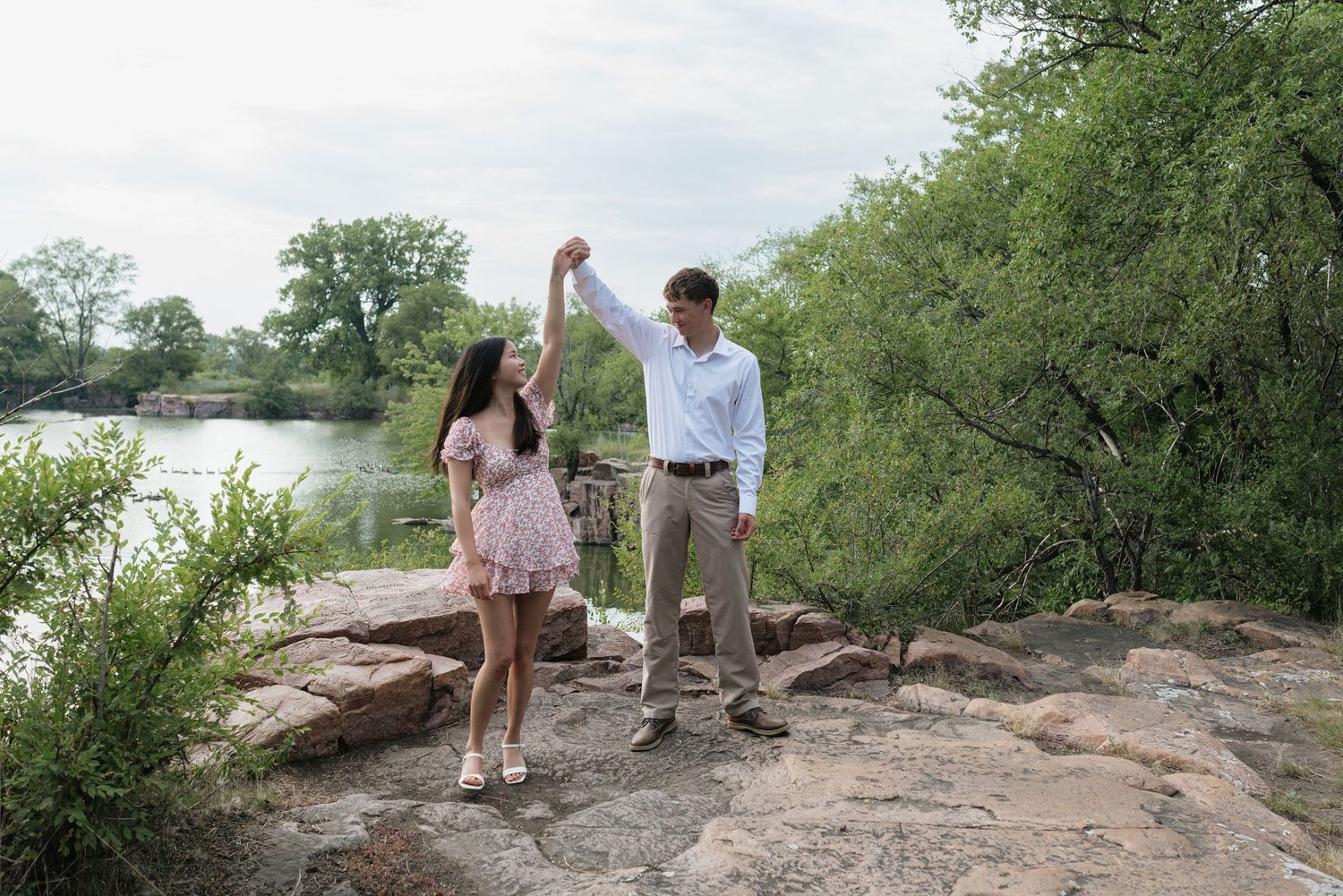 Couple dancing on rocky terrain near a lake surrounded by green trees on a sunny day.