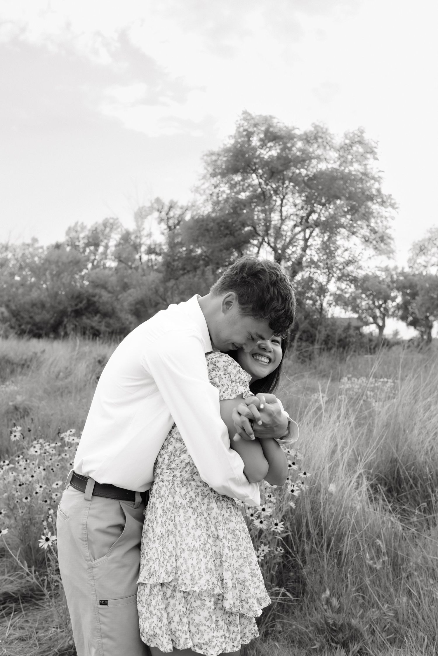 Black and white photo of a laughing couple hugging in a wildflower field, man in white shirt and woman in floral dress.