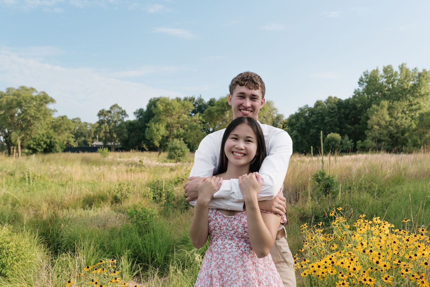 A smiling couple posing together in a sunny meadow with yellow wildflowers and lush greenery.