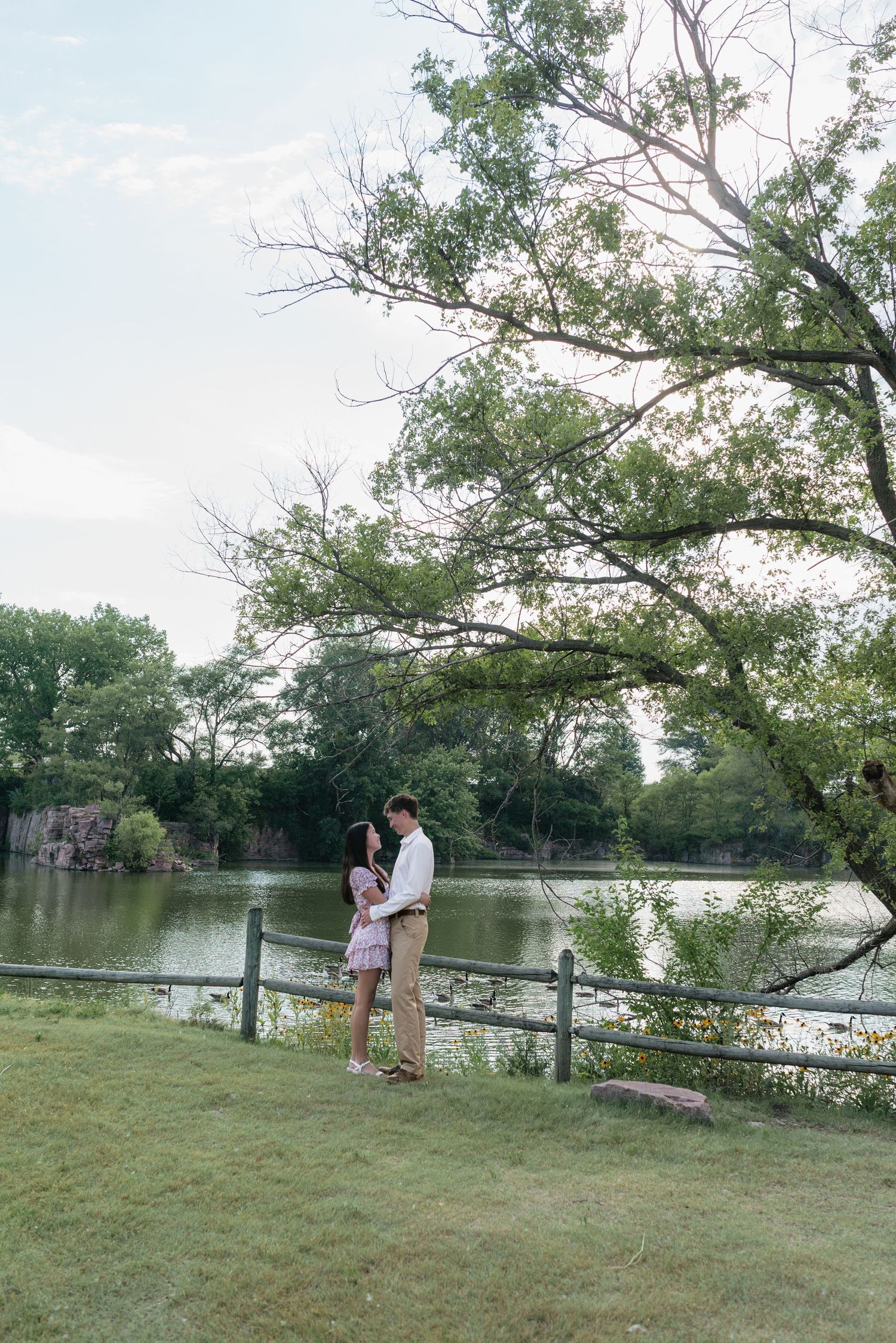 Couple standing together by a serene lake with lush green trees and a wooden fence in a park.